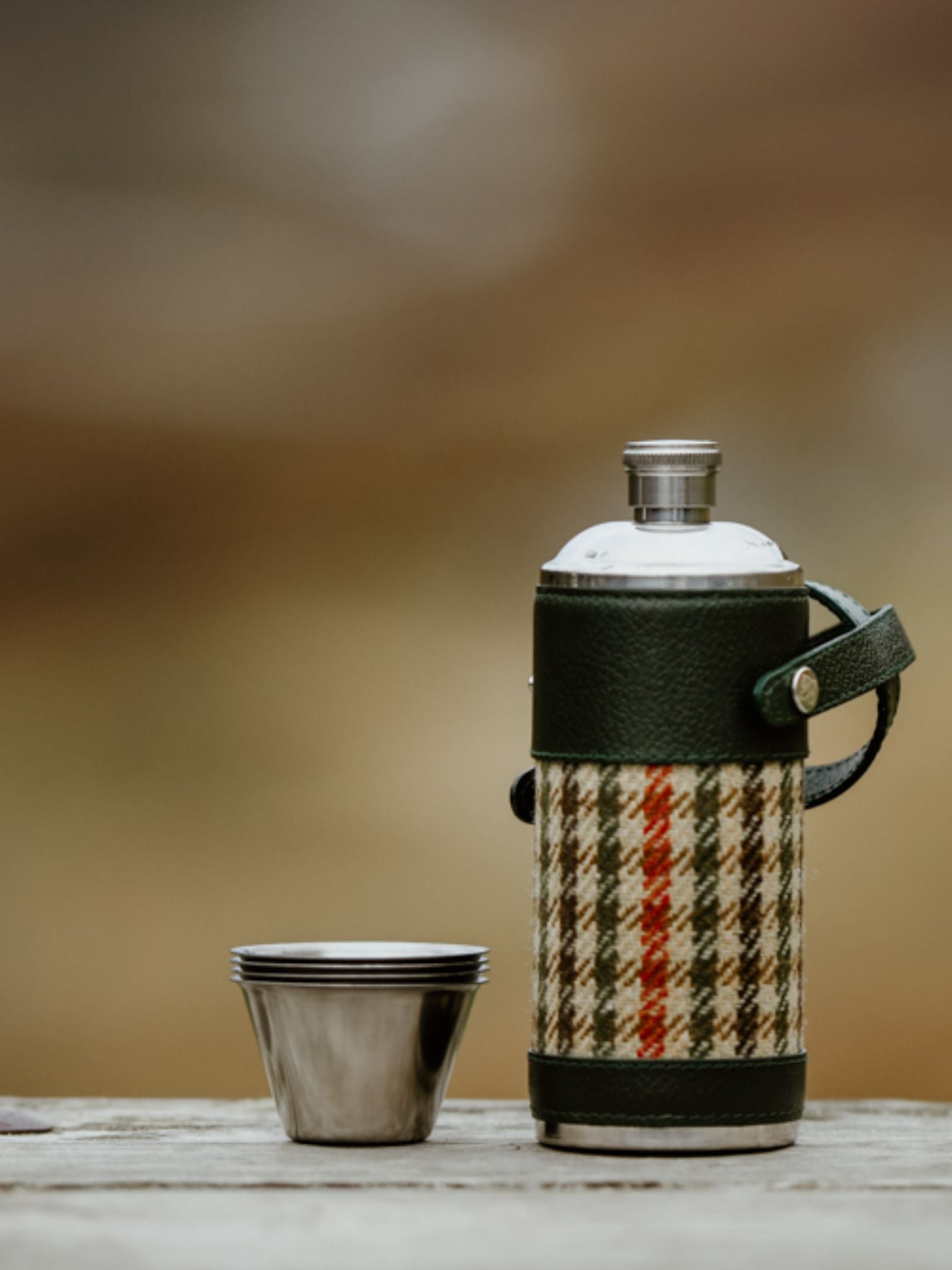 The 8oz Bordeaux Hunter Flask by Campbell’s of Beauly x Ettinger, with a green plaid cover and Capra goat leather strap, stands next to three stacked stainless steel cups on wood, set against a blurred outdoor backdrop—exuding British craftsmanship.