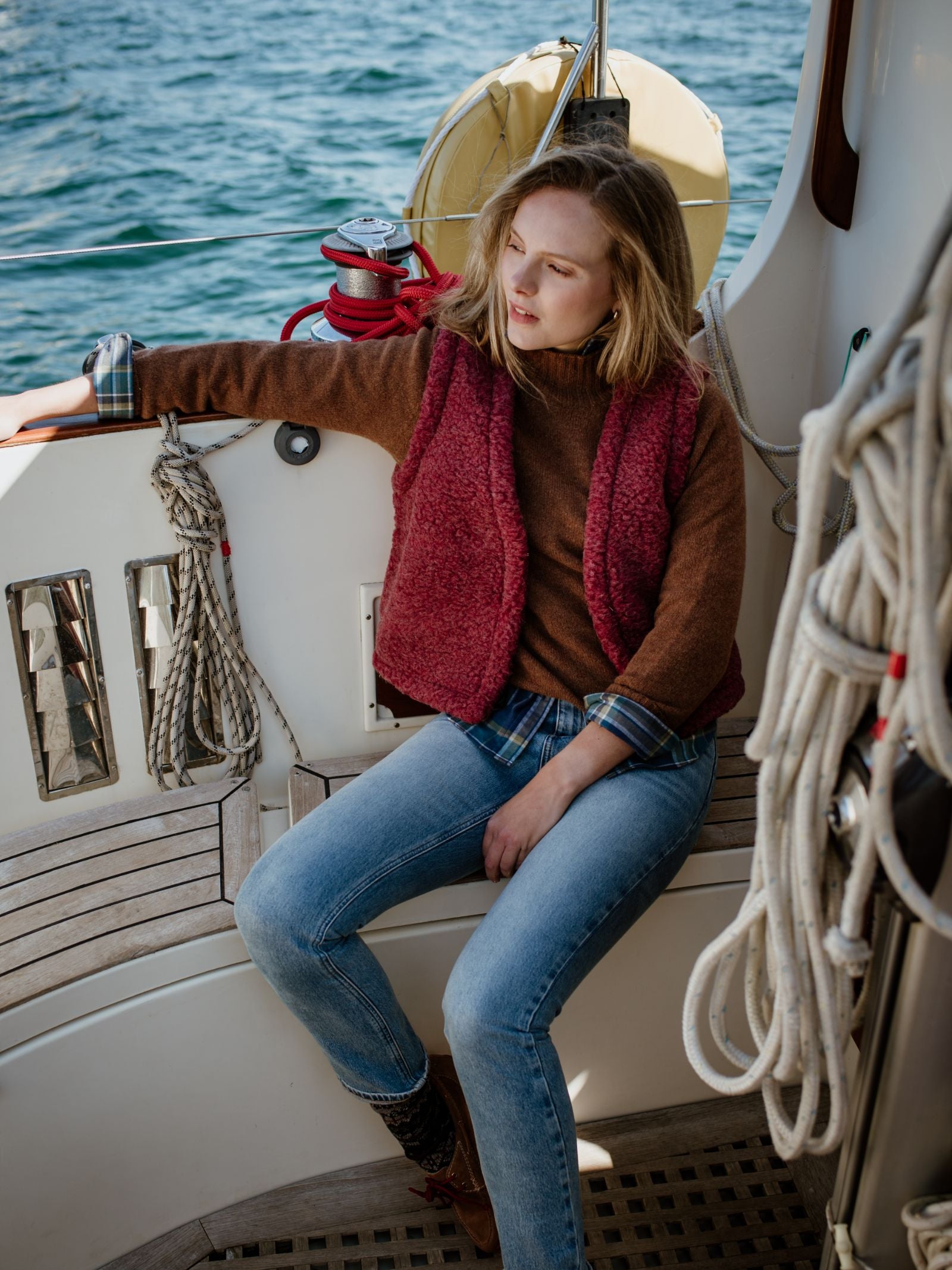 A woman in a maroon Campbell's of Beauly Wool Shrug and jeans relaxes on a sailboat, enjoying its lightweight warmth and easy layering. Nautical ropes, wooden deck, and blue water complete the scene.