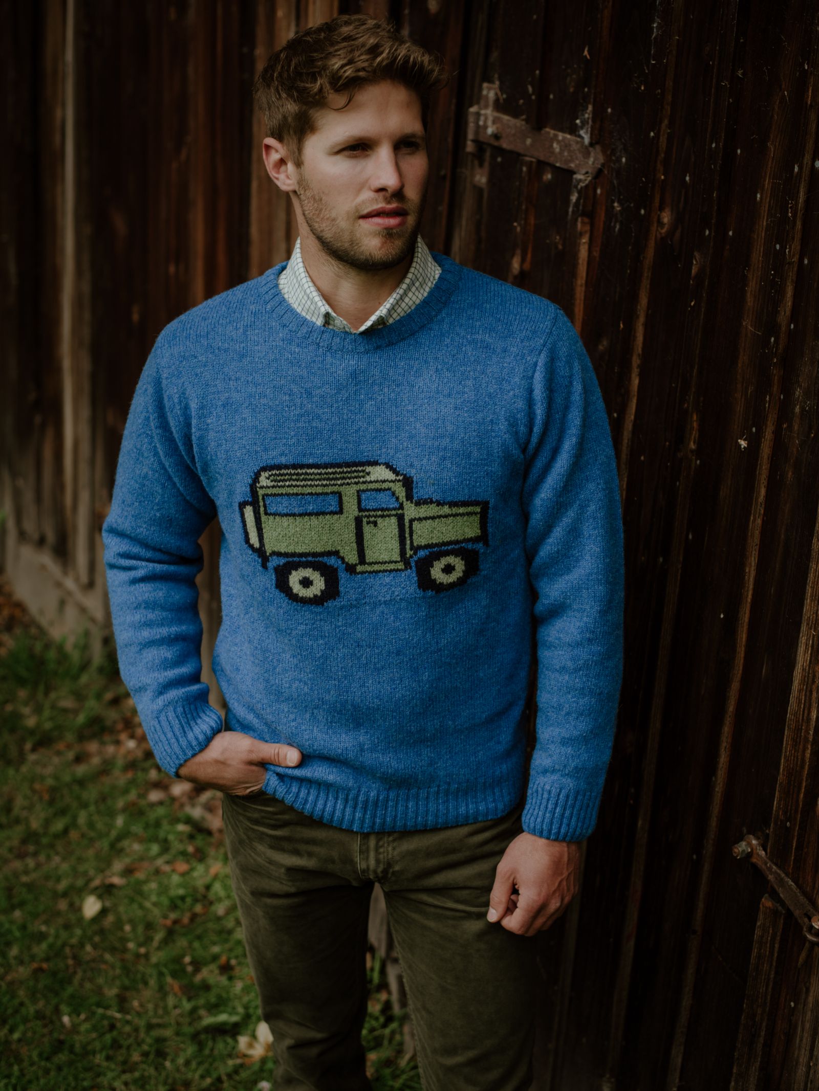 A man with light brown hair and a short beard stands outside by a wooden wall, wearing the Campbell's of Beauly Unisex Land Rover Defender Jumper—a classic knitwear blue sweater with a green and black car graphic—plus an olive-green shirt and pants.