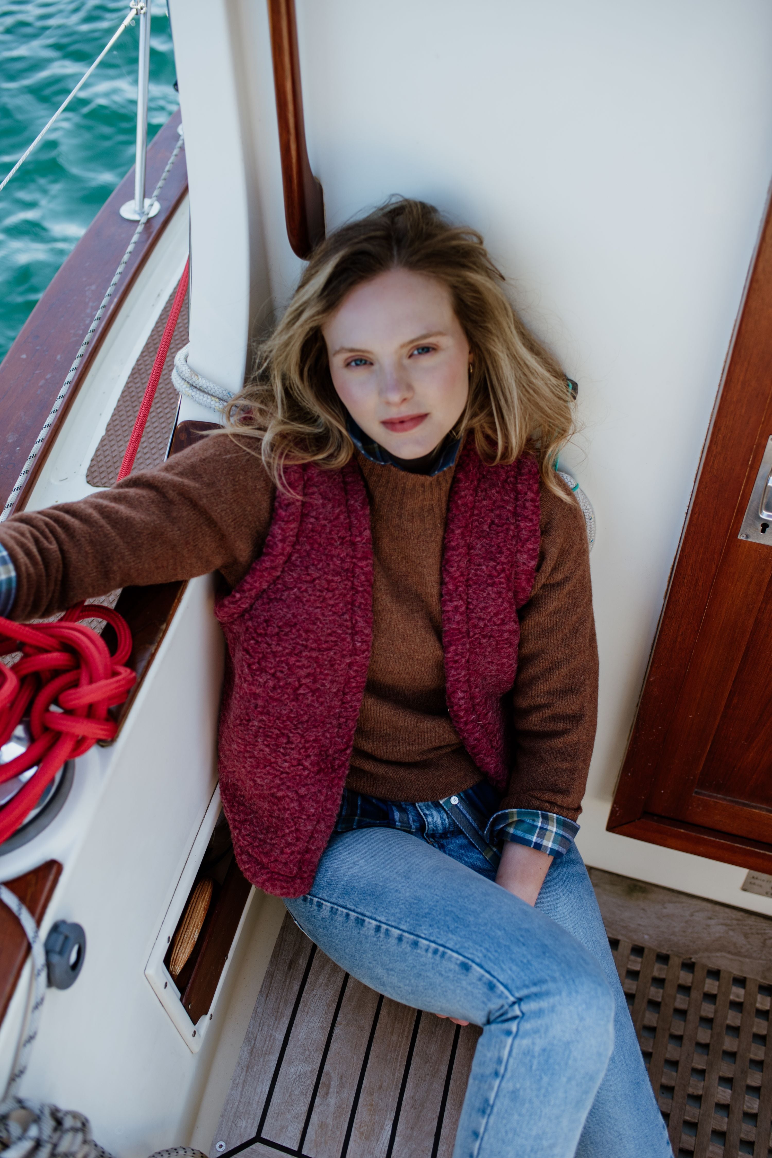 A woman with long blonde hair sits on the wooden floor of a boat, leaning against the white wall. She wears a brown sweater, a red vest, blue jeans, and looks calmly at the camera. Red rope is beside her.