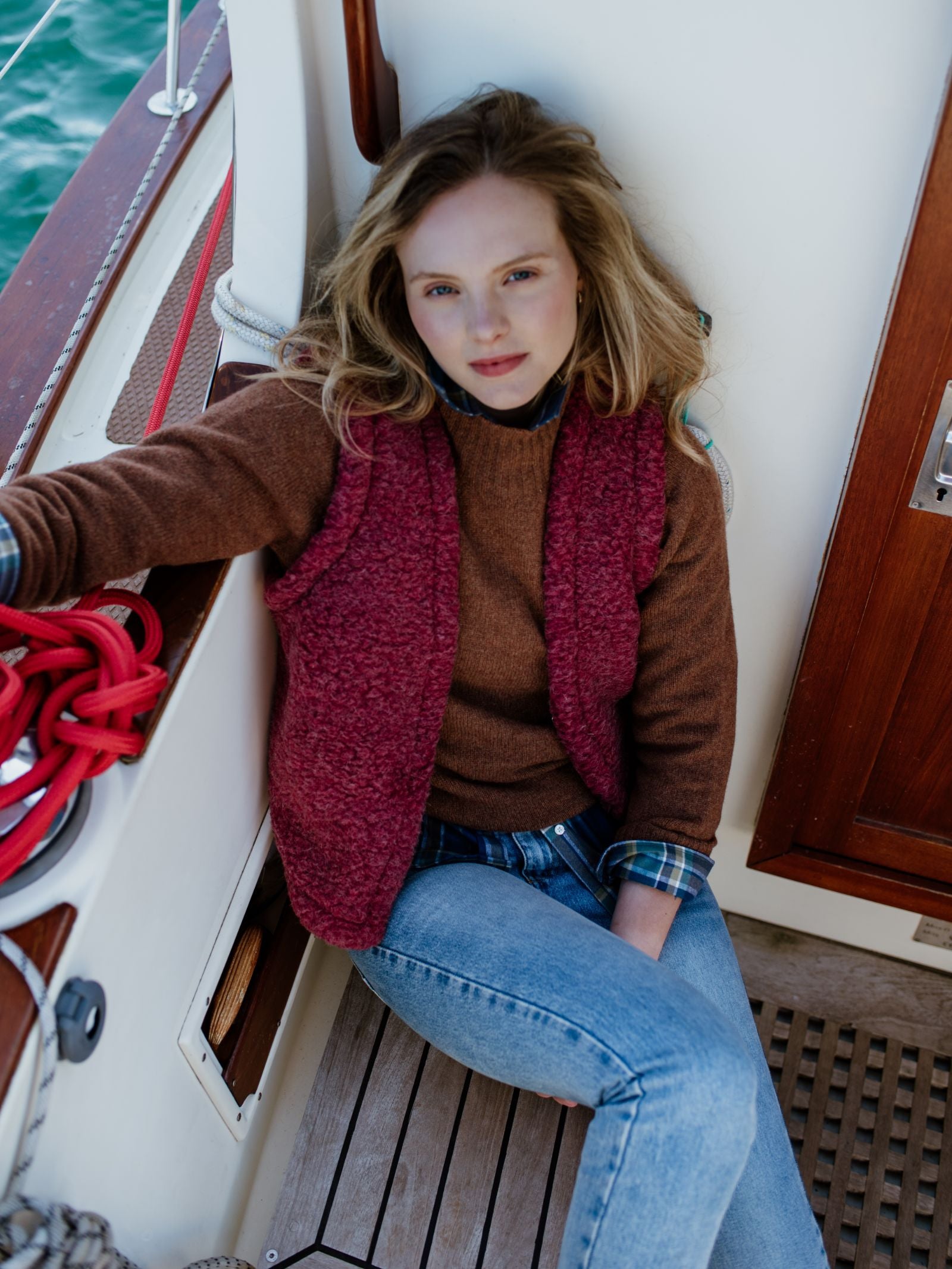 A woman with light brown hair sits on a boat, leaning against the wall in a Campbell's of Beauly Wool Shrug layered beneath a red vest and paired with blue jeans. Ropes and wooden elements add to the relaxed nautical setting.