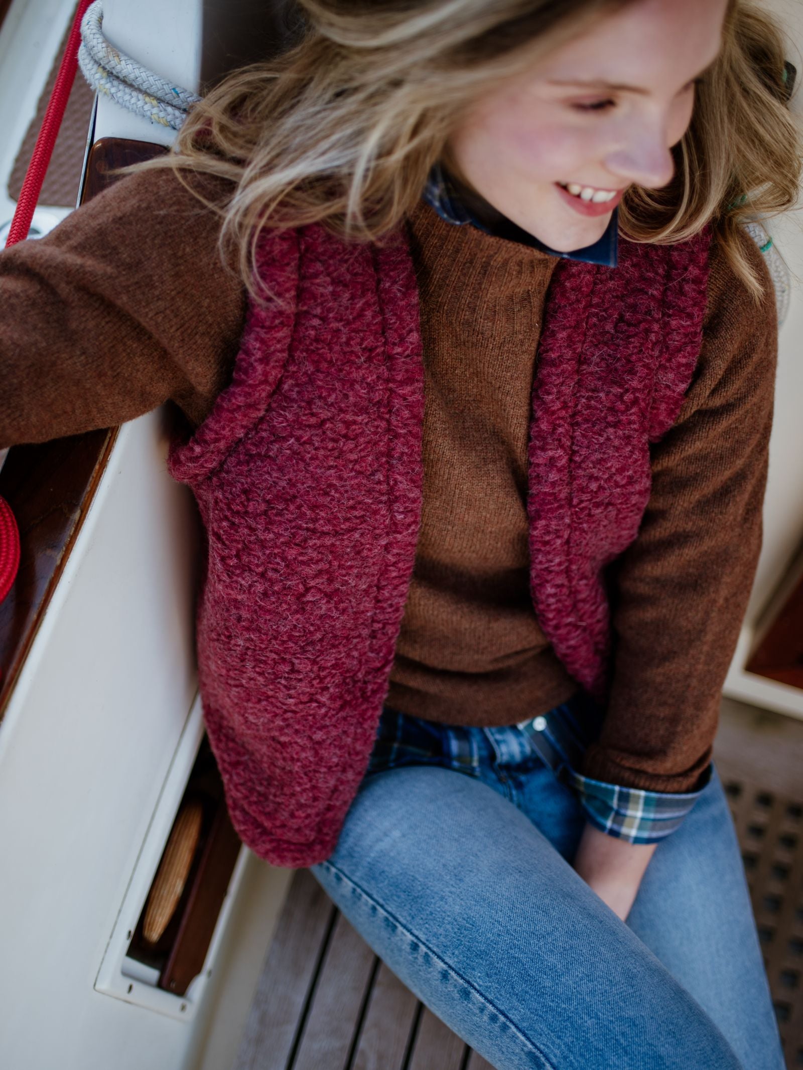 A smiling woman with long blonde hair sits on a boat, wearing the Campbell's of Beauly Wool Shrug layered over a blue plaid shirt and light blue jeans.