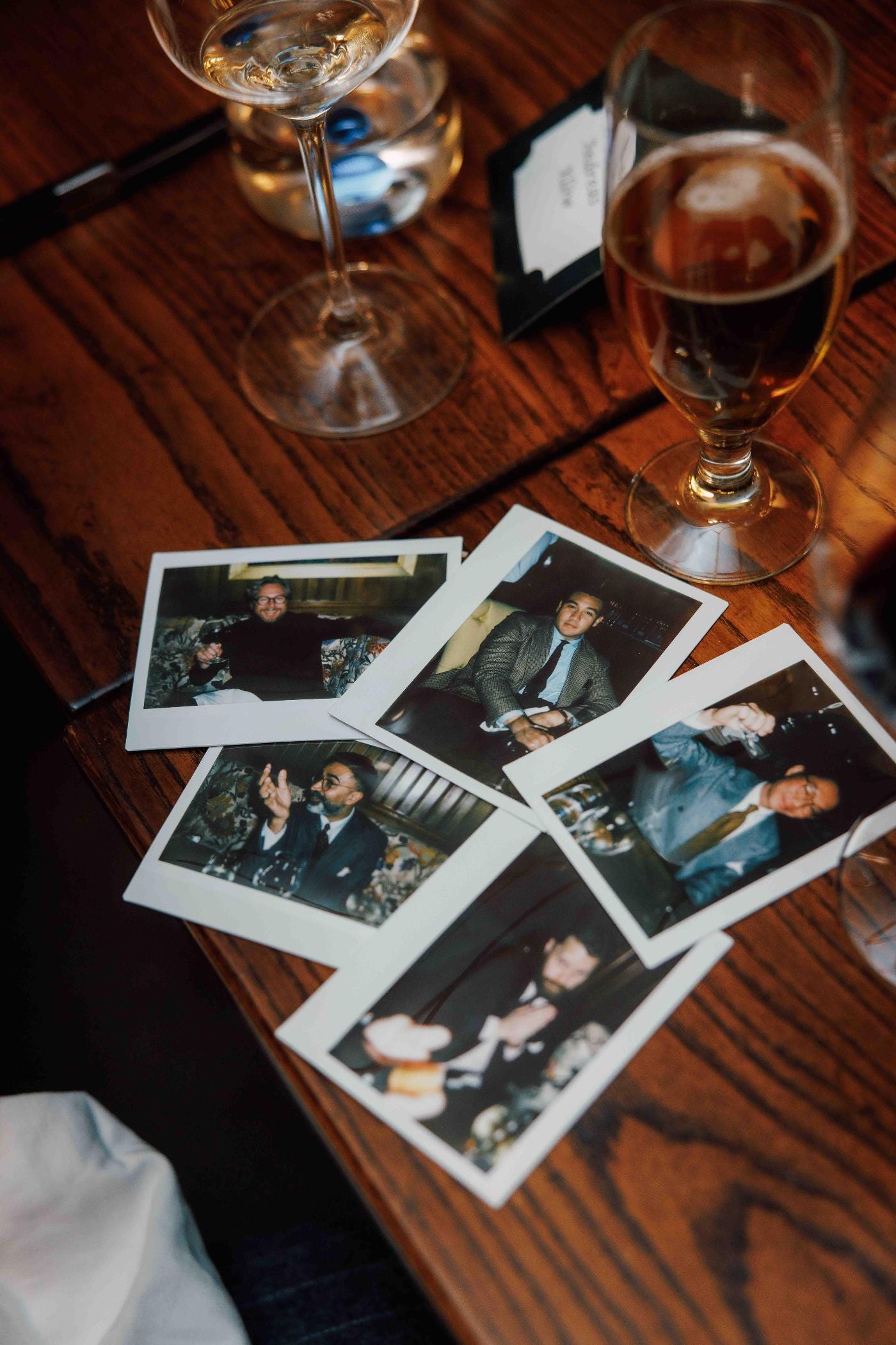 Six instant photos showing people enjoying drinks are spread out on a wooden table, surrounded by glasses of wine, beer, and water. The scene suggests a lively social gathering.