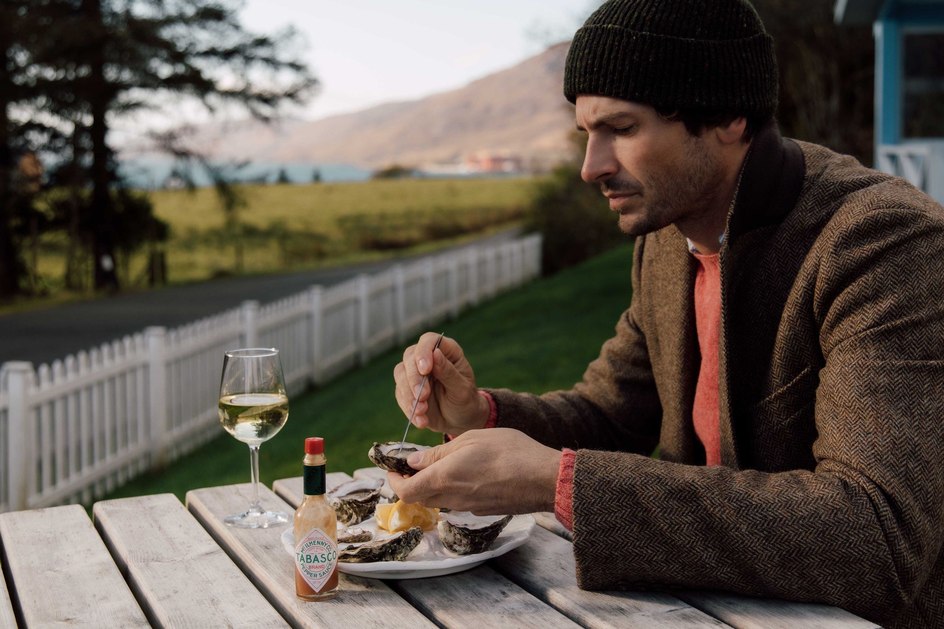 A man in a jacket and beanie sits at an outdoor table, eating oysters with lemon. A glass of white wine and a bottle of Tabasco sauce are beside him. There’s a white fence, green lawn, and mountains in the background.