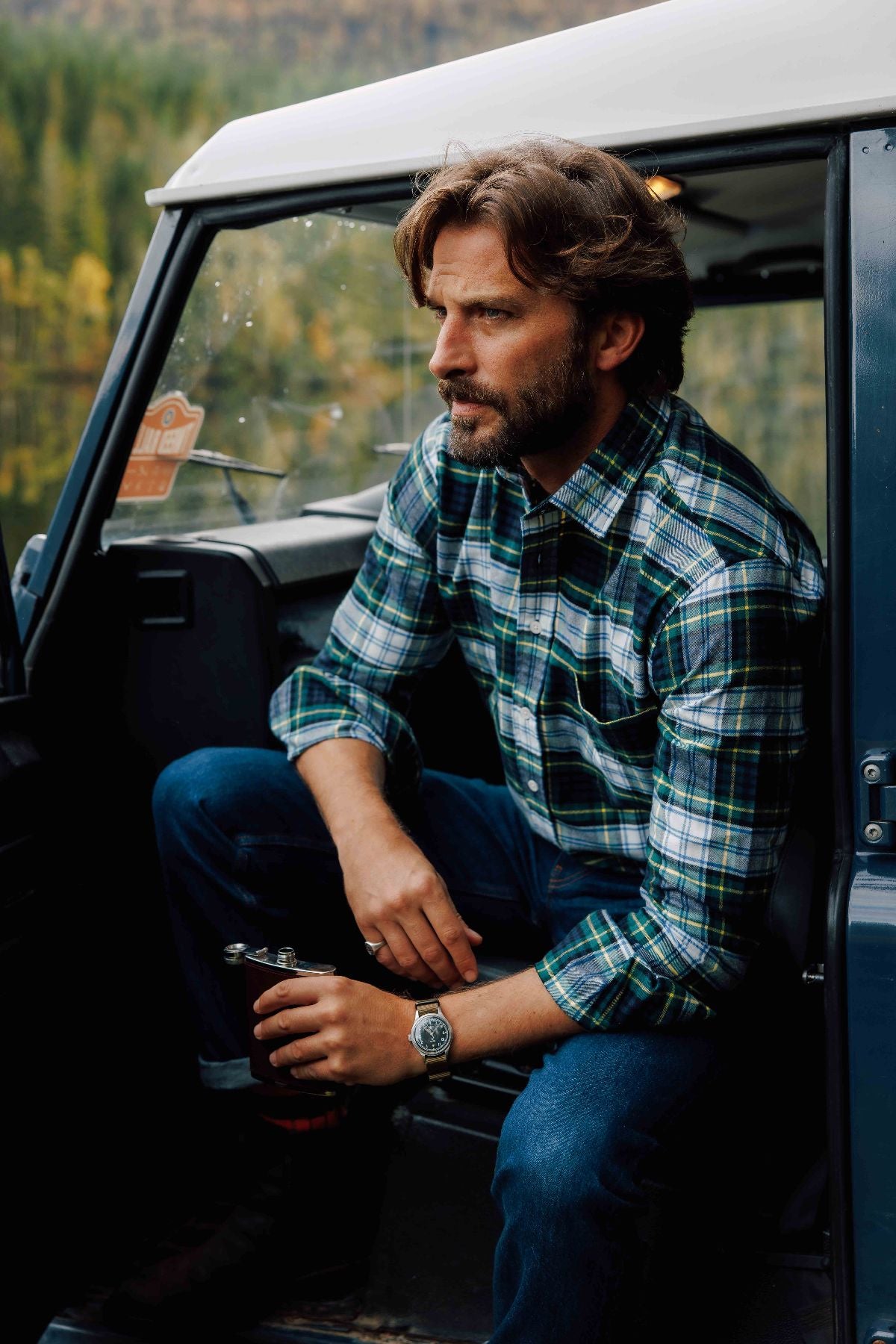 A bearded man in a green plaid shirt and jeans sits on the open door frame of a blue vehicle, holding a flask and looking thoughtfully into the distance. A forest scene is visible in the background.