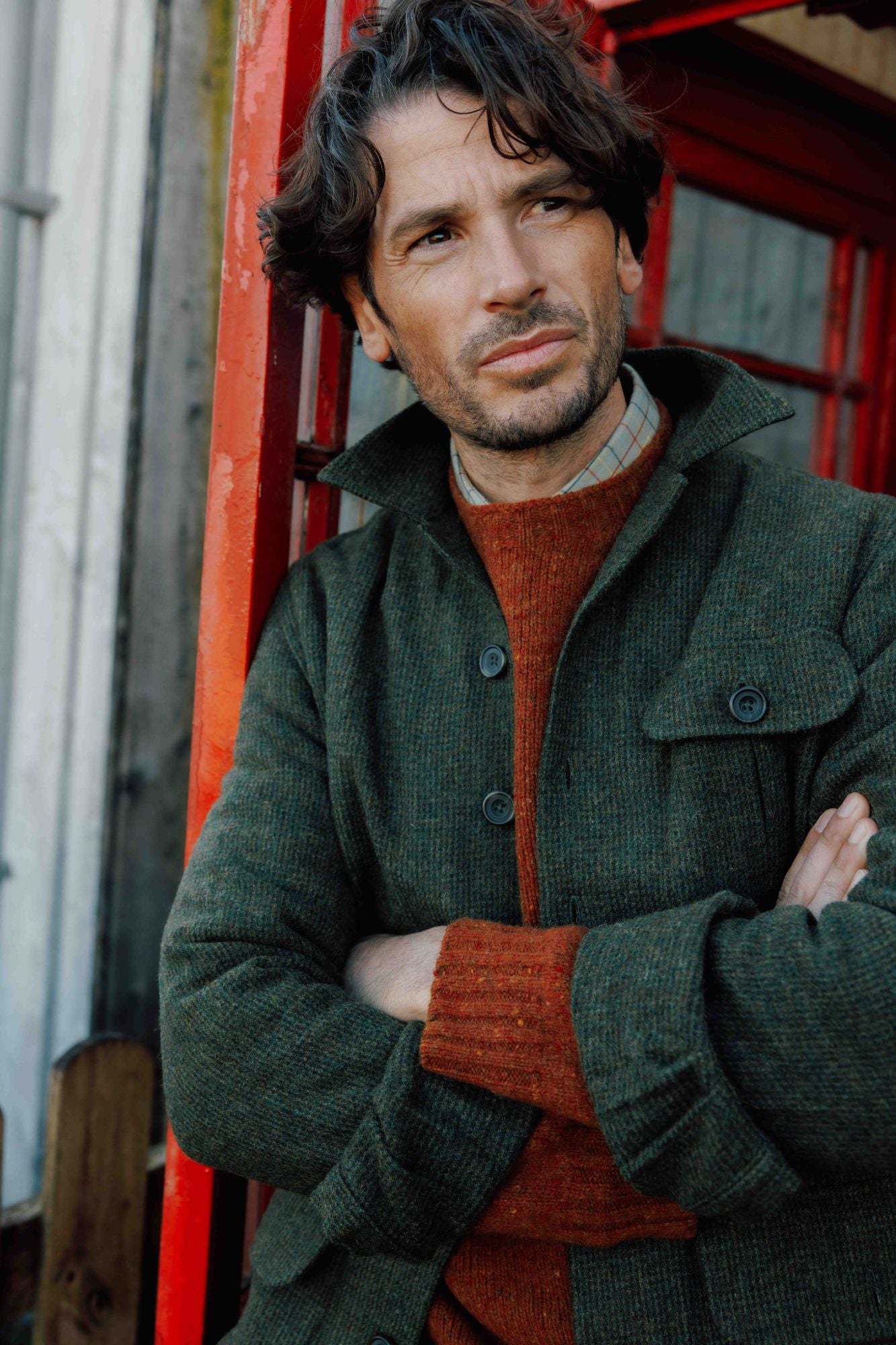 A man with wavy dark hair and stubble stands with crossed arms, wearing a green coat over a rust-colored sweater, in front of a bright red phone booth. He looks thoughtfully into the distance.