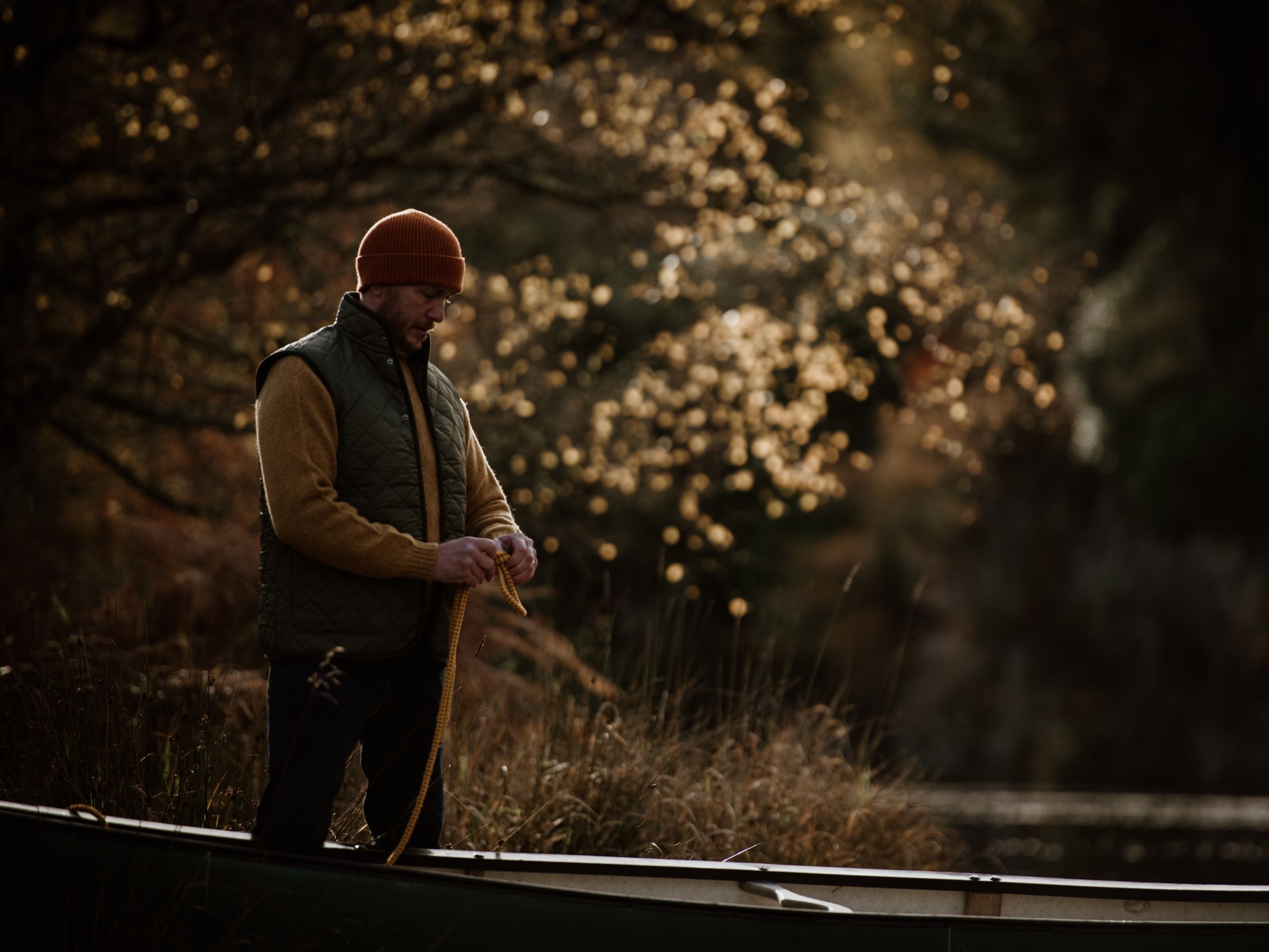 A man in a beanie and vest stands beside a canoe, holding a rope. Autumn trees with golden leaves and soft sunlight create a warm, serene atmosphere in the background.
