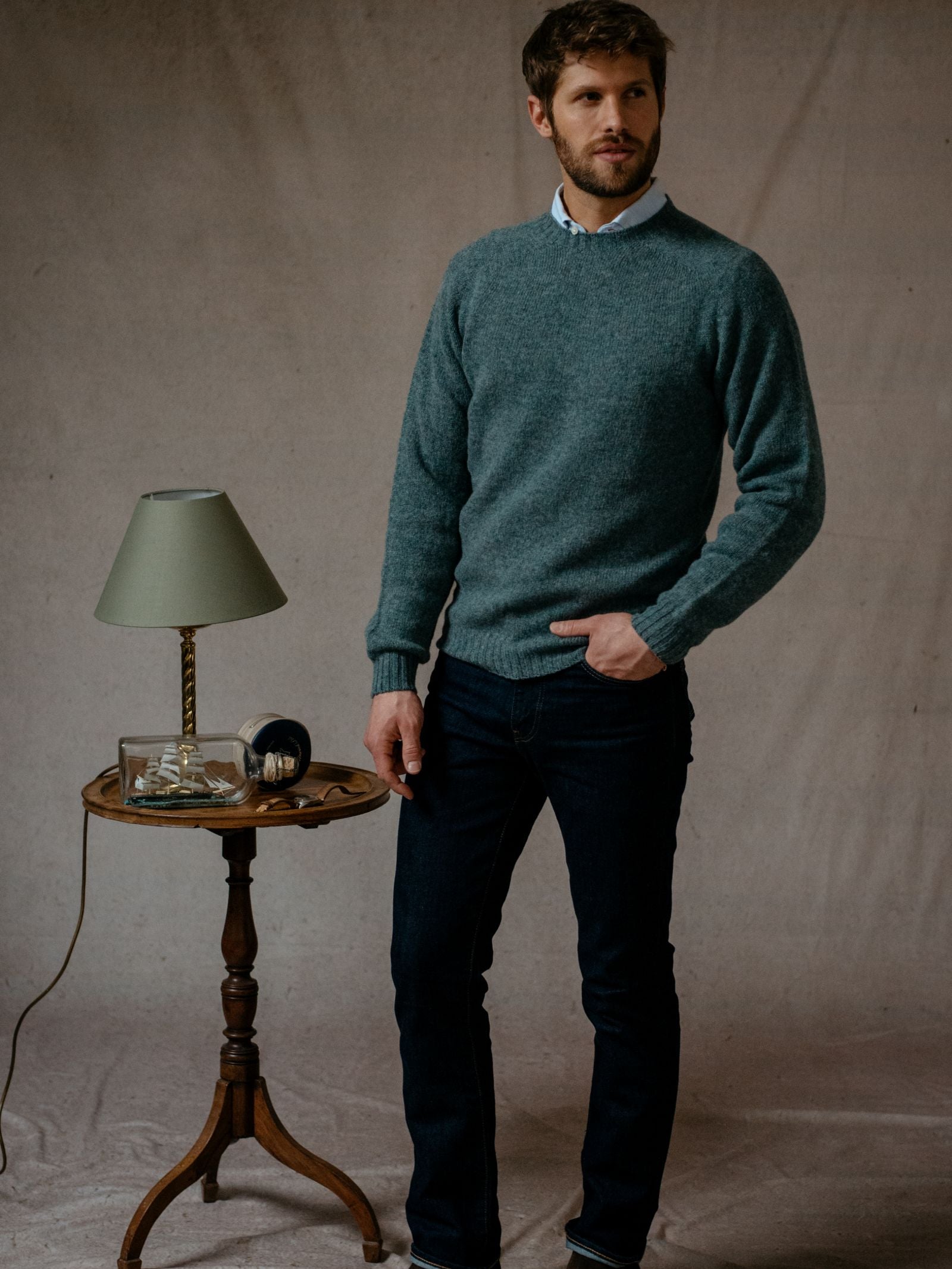 A man with short brown hair and a beard stands beside a small round table. He wears the Shetland Jumper by Campbell's of Beauly over a collared shirt and dark jeans, one hand in his pocket.