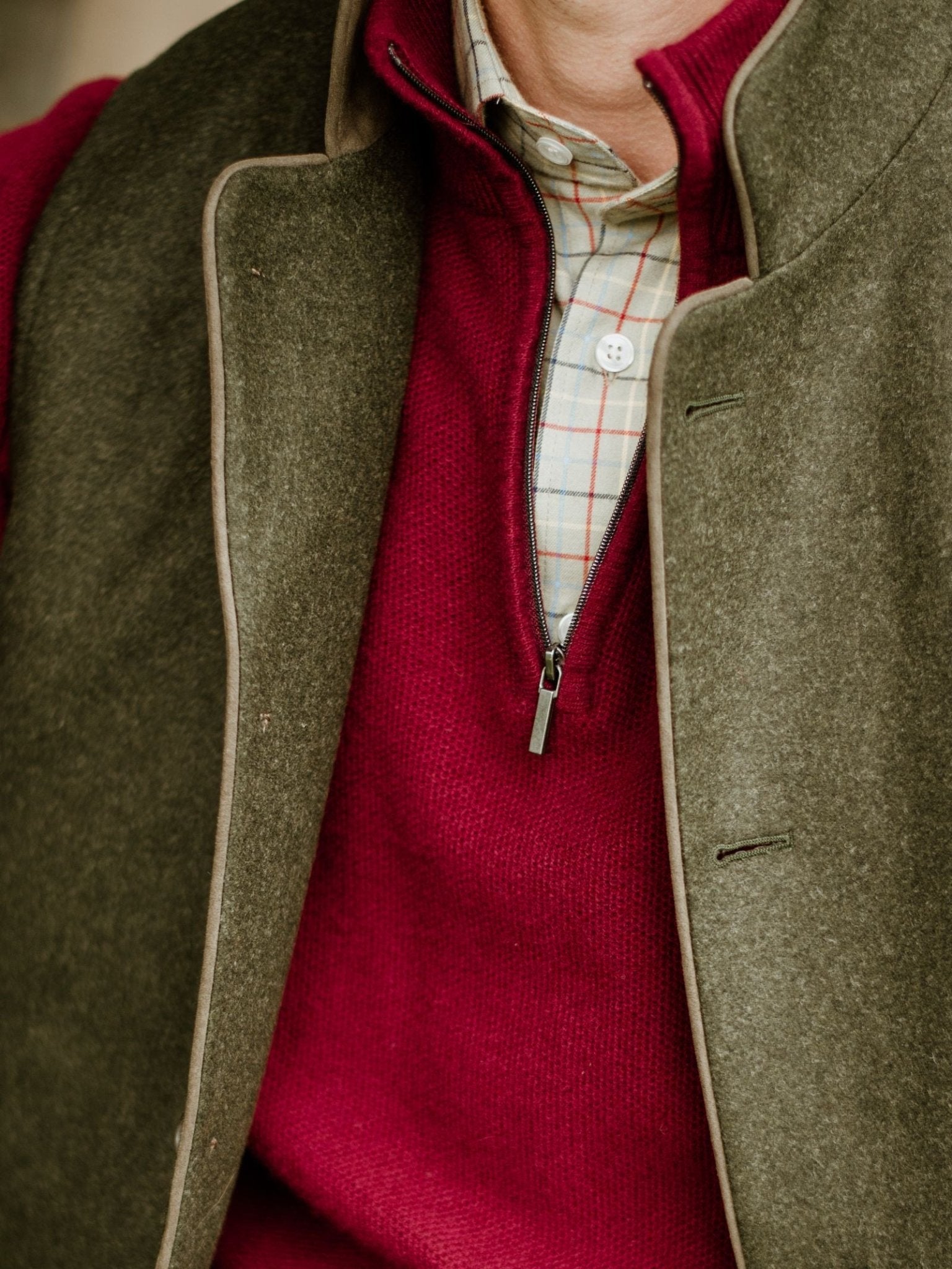 A person models the Campbell's of Beauly Luxury Field Nehru Gilet in Loden green, layered over a red sweater and light plaid shirt. The photo highlights the upper torso and neck, keeping the face unseen.