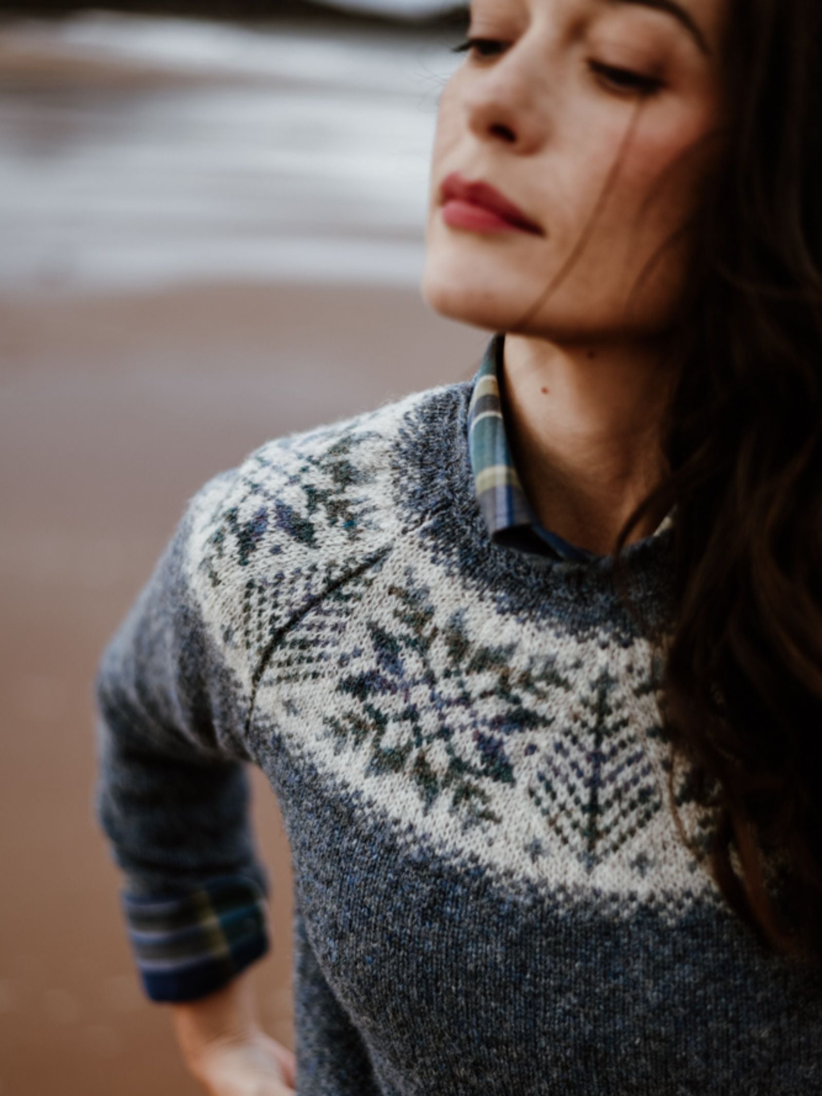 A woman stands outdoors on a blurred sandy beach, wearing Campbell's of Beauly's Auld Stock Shetland Wool Snowflake Crew Jumper over a plaid shirt. She looks slightly downward, her hair draped to one side.