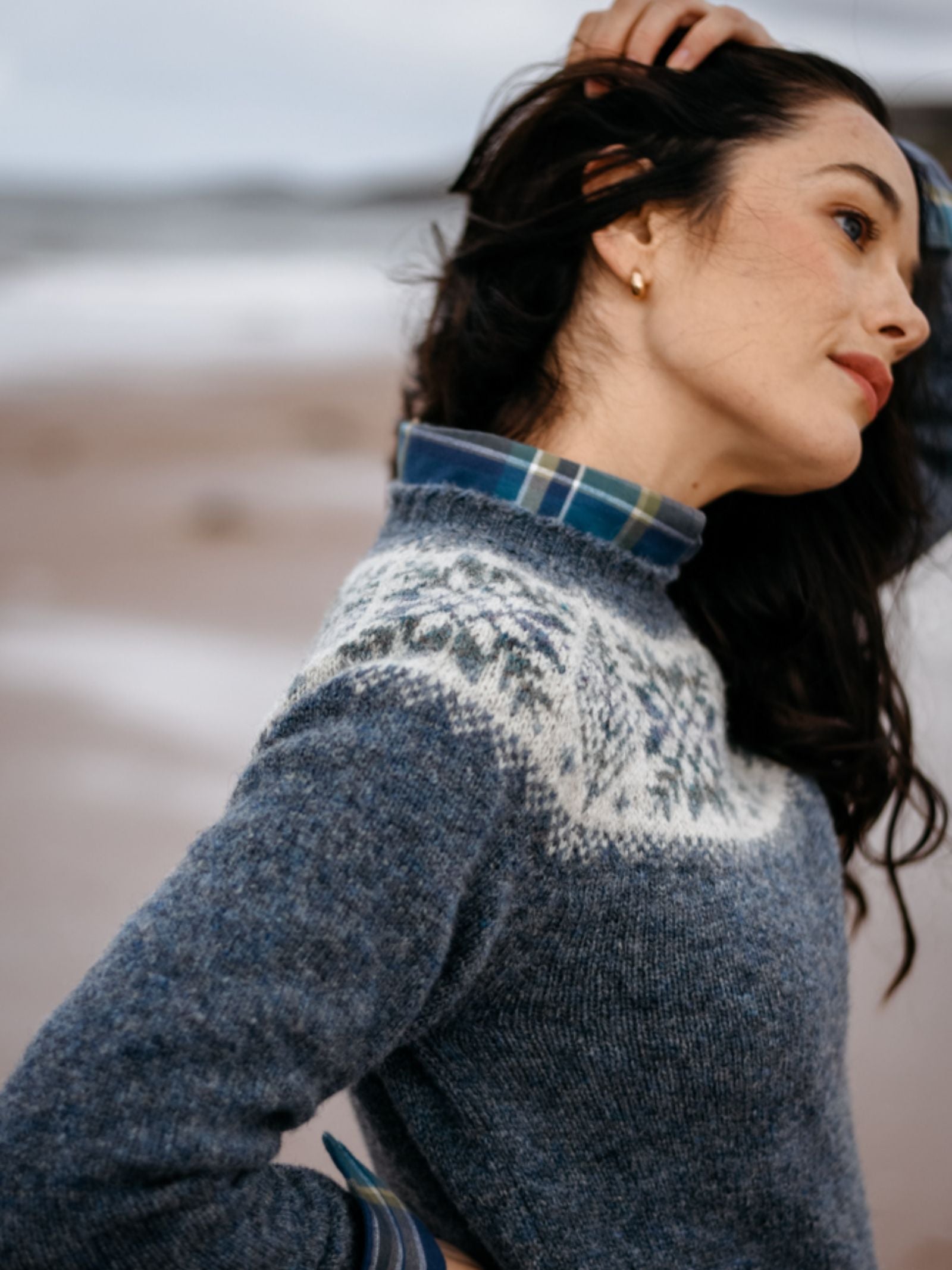 A dark-haired woman poses sideways at the beach, thoughtfully touching her hair while wearing Campbell's of Beauly's Auld Stock Shetland Wool Snowflake Crew Jumper over a plaid shirt, embodying cozy, timeless winter style.