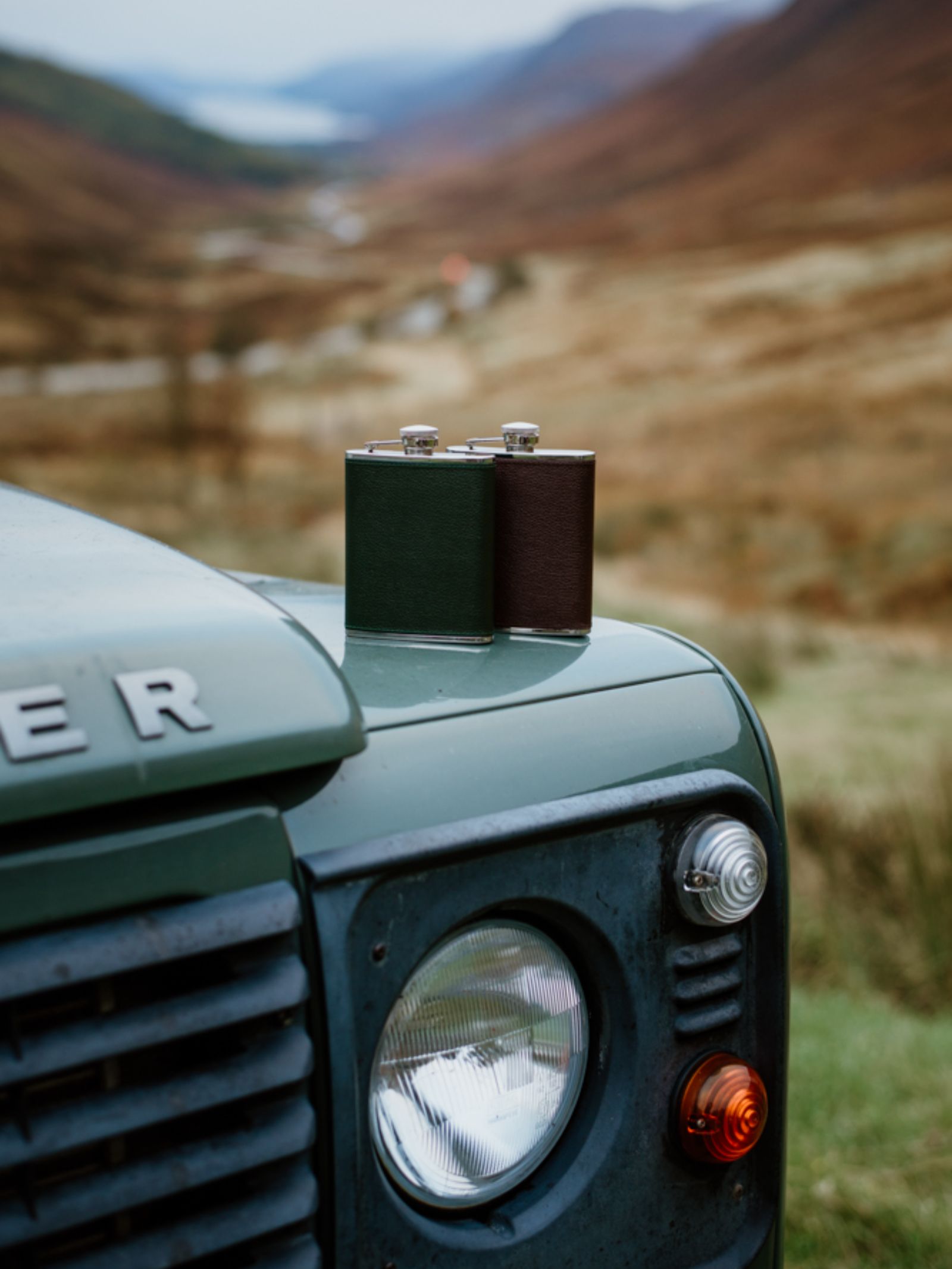 A 6oz Forest Green Hip Flask by Campbell's of Beauly x Ettinger rests on a green Land Rover’s hood, parked amid mountains and a winding road—celebrating British craftsmanship and the spirit of adventure.