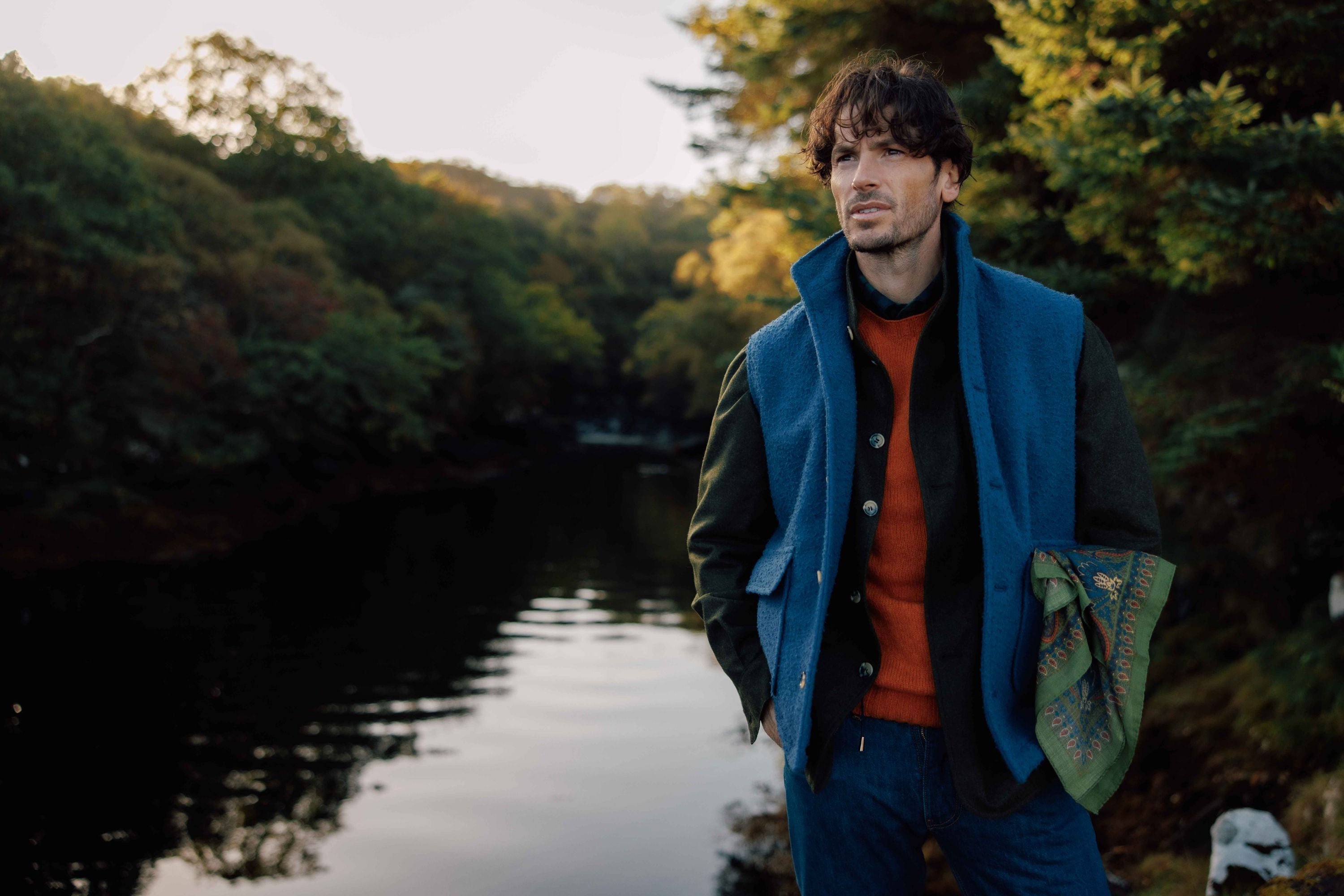 A man stands by a calm river surrounded by trees, wearing a blue vest, orange sweater, dark jacket, and blue pants, holding a patterned green scarf, with sunlight filtering through the foliage.