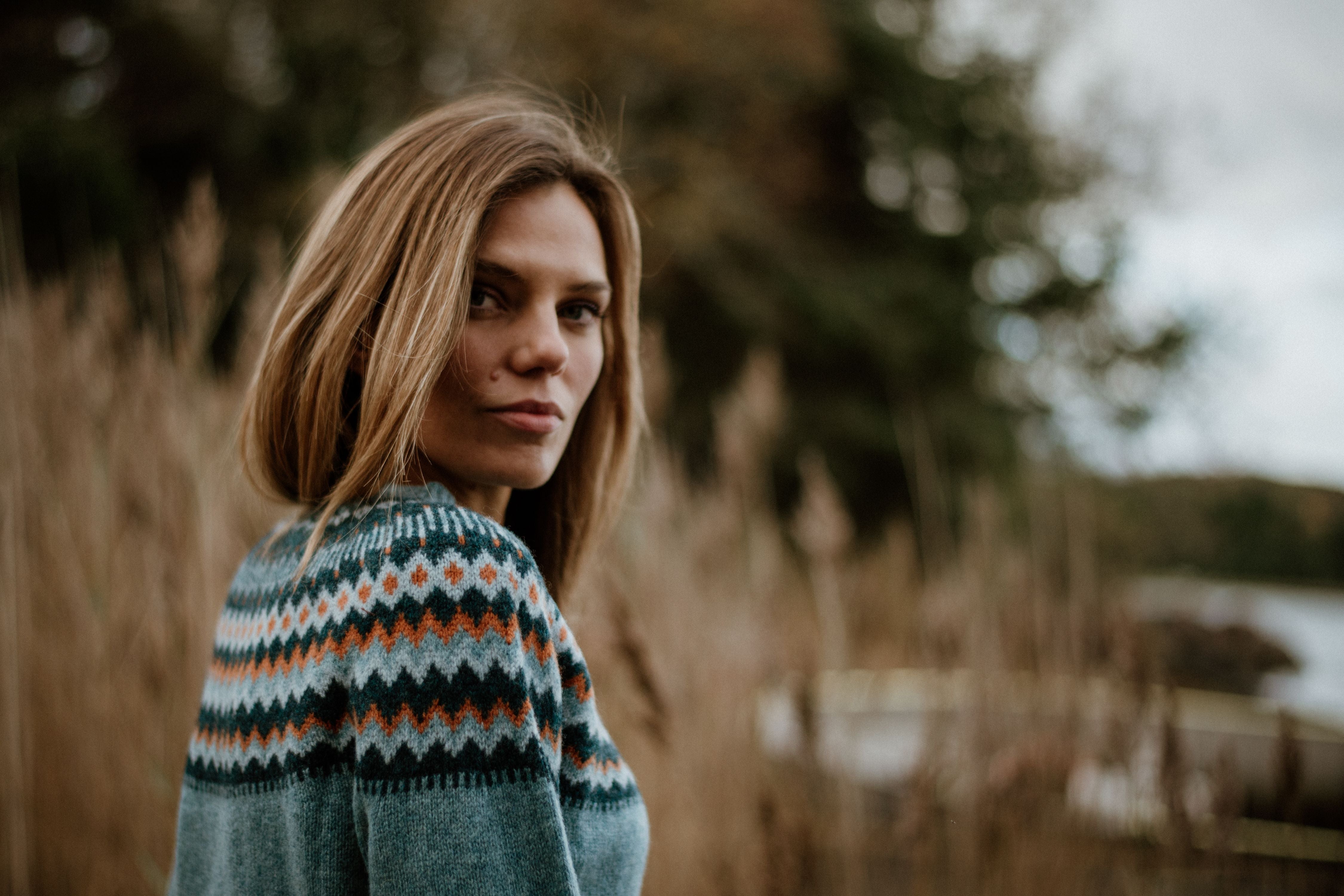 A woman with straight, light brown hair wears a patterned sweater and stands outdoors in front of tall grasses and blurred trees, looking back over her shoulder at the camera.