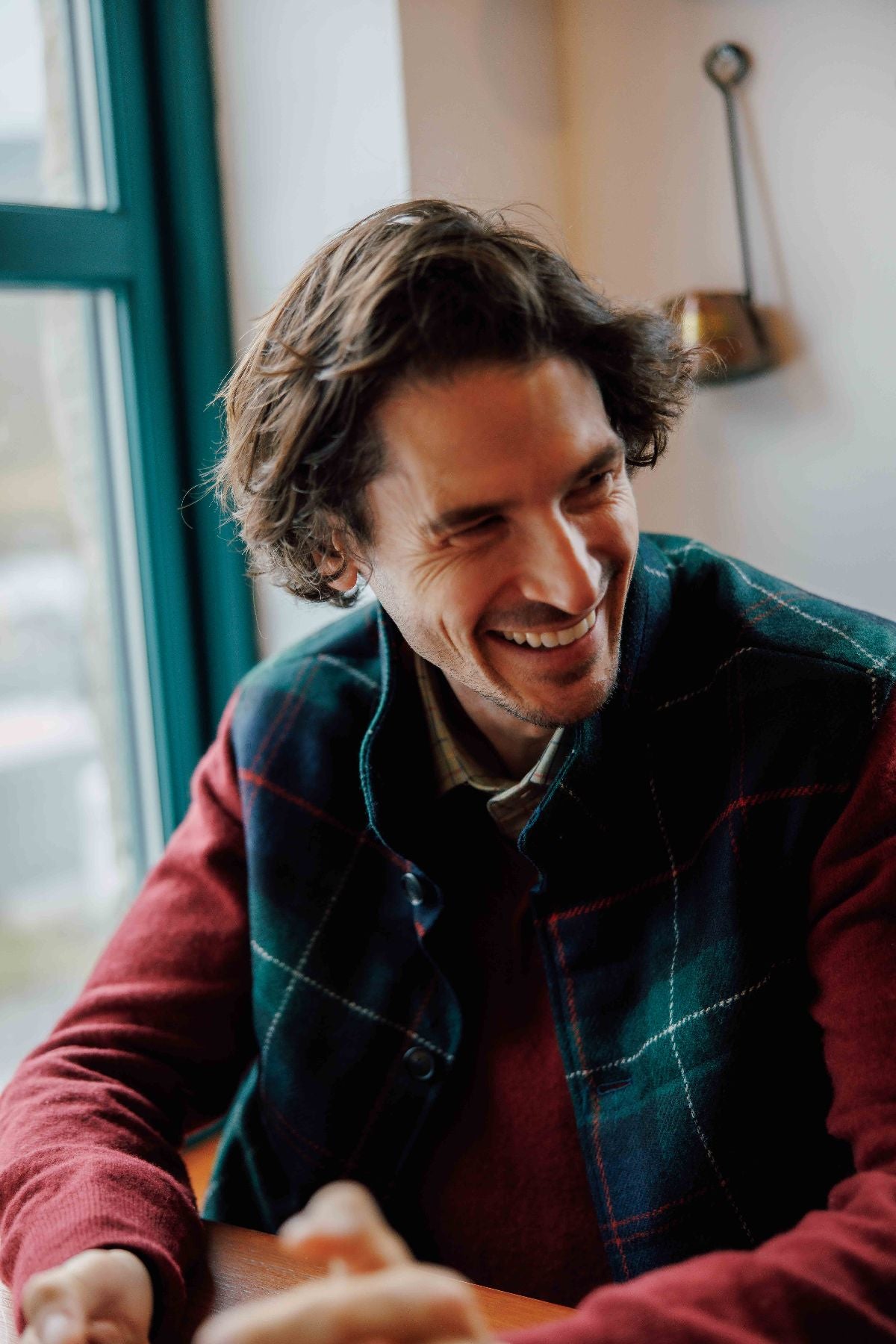 A man with wavy brown hair and a trimmed beard smiles while sitting indoors by a window, wearing a red sweater and a plaid vest. Warm light creates a cozy atmosphere.