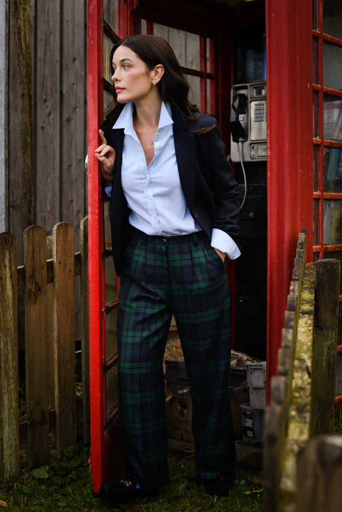 A woman wearing a blue button-up shirt, dark blazer, and green plaid trousers stands in the doorway of a red British phone booth, looking to the side with hands in her pockets. A wooden fence and building are in the background.