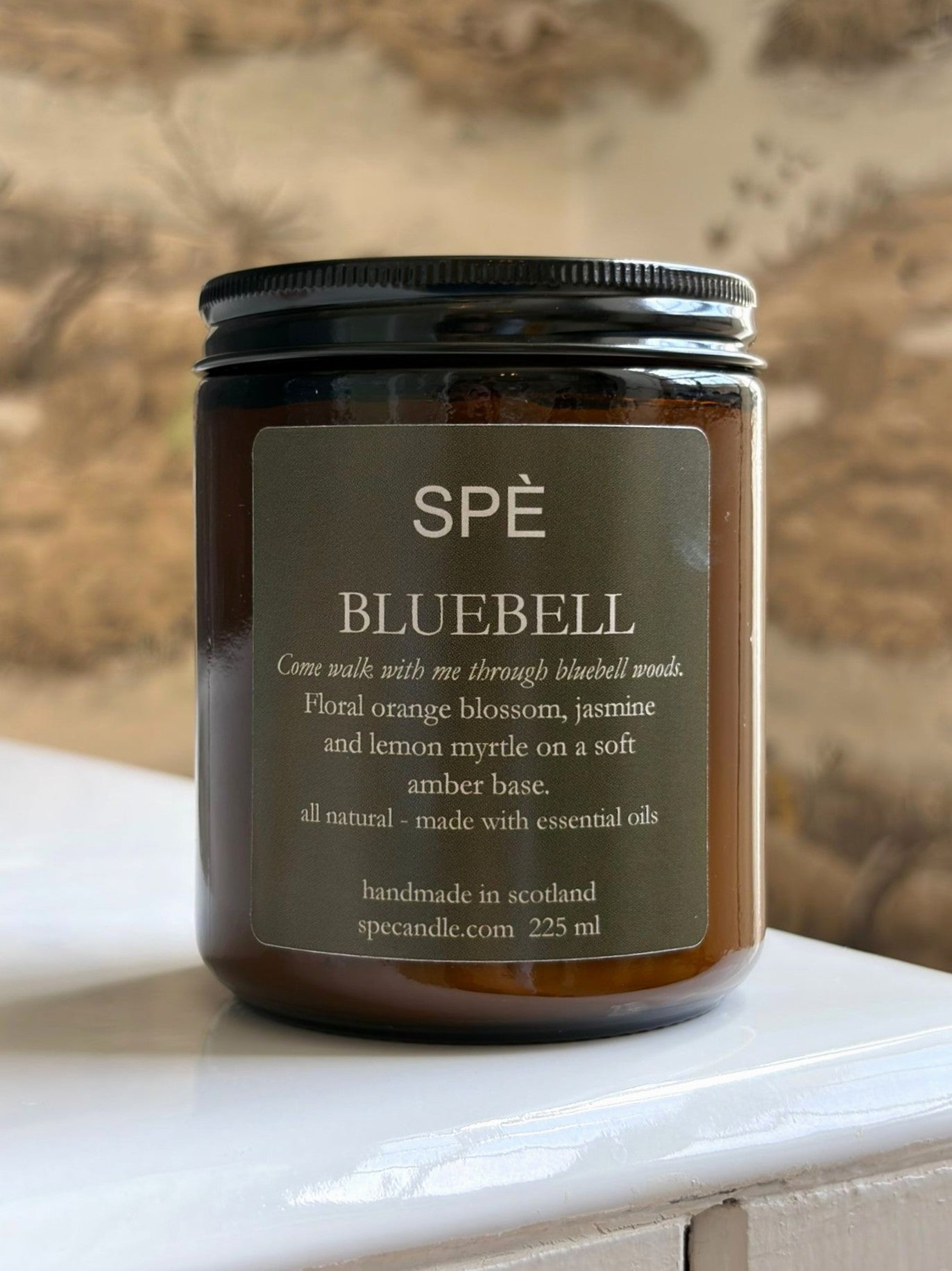 A brown glass Bluebell Candle by SPÈ Atelier, featuring a label that reads “SPĒ BLUEBELL.” Handmade in Scotland, this floral candle offers scent notes of orange blossom, jasmine, lemon myrtle, and amber. It is displayed on a white surface.