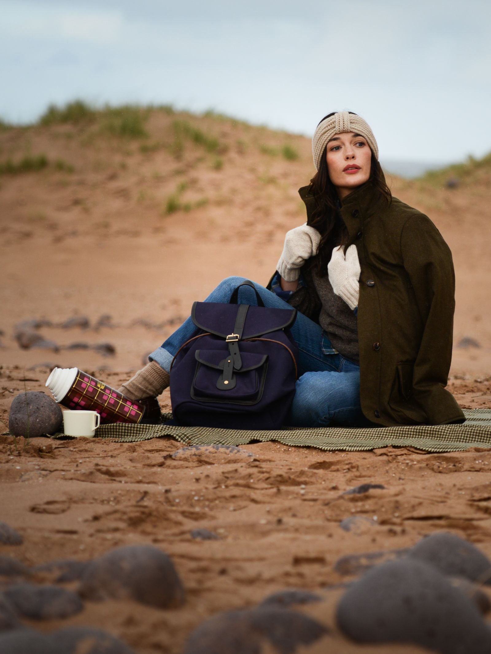 A woman wearing Campbell's of Beauly Chunky Mittens, a beanie, and a coat sits on a blanket at the beach with her backpack, thermos, and cup beside her, looking to the side with rocks and sand in the background.
