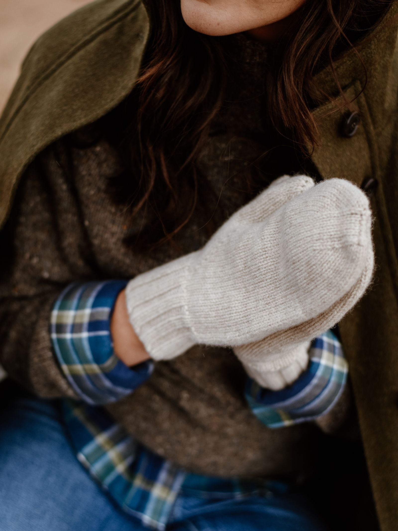 Wearing a green coat, brown sweater, and plaid shirt, this person holds their hands together in Chunky Mittens by Campbell’s of Beauly—a thick white lambswool accessory made from natural fibres. Only the lower face and torso are shown.