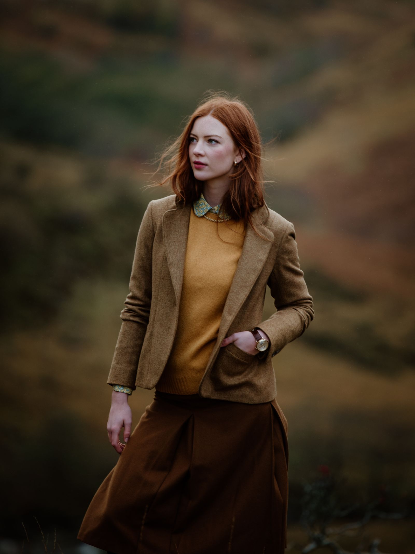 A woman with long red hair stands outdoors in the Auld Stock House Jacket by Campbell's of Beauly, layered over a mustard sweater and brown skirt against a blurred natural backdrop.