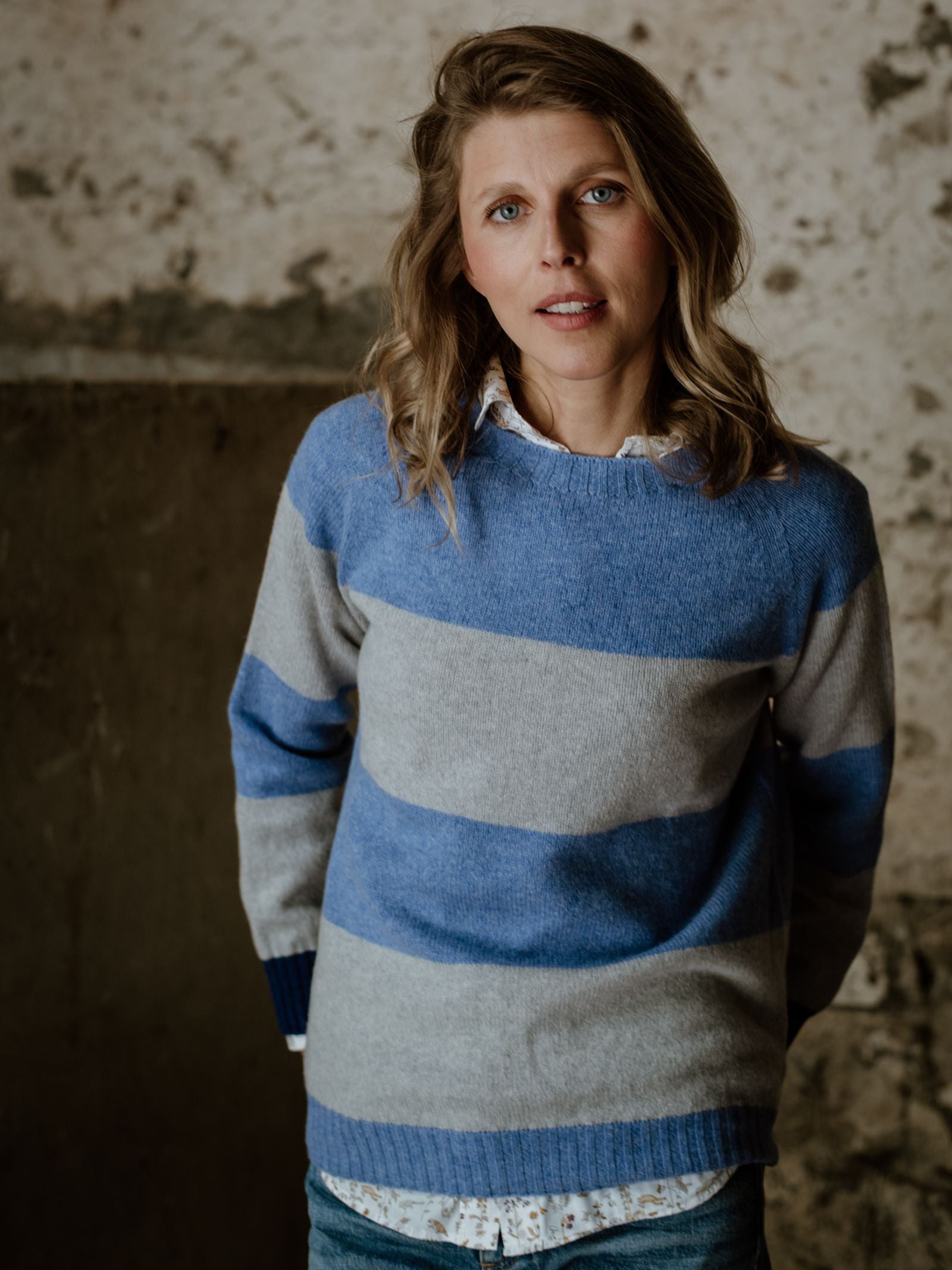A woman with wavy blonde hair wears the Campbell's of Beauly Auld Stock Stripe Crew Jumper, a blue and gray striped knit made in Scotland, standing calmly before a rustic textured wall and looking at the camera.
