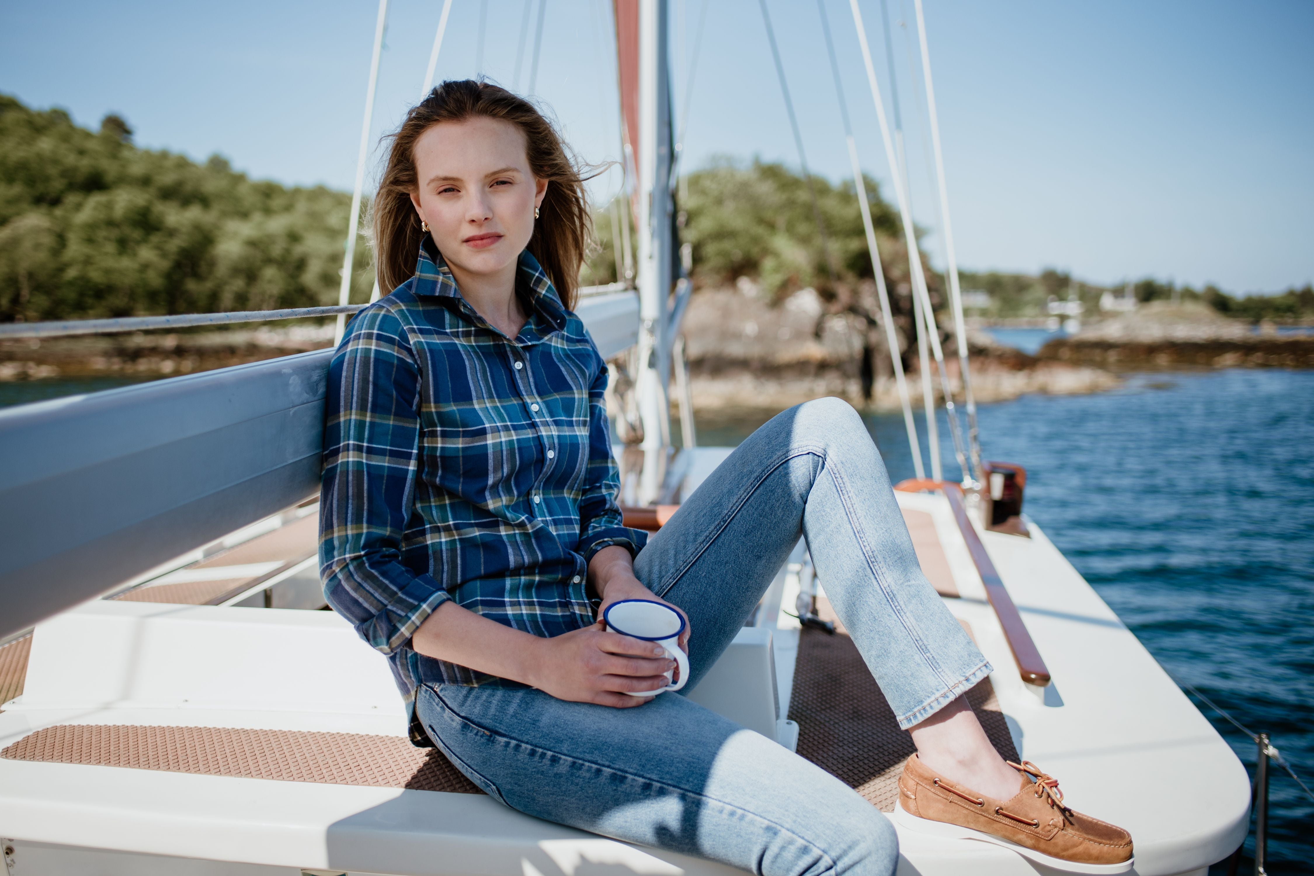 A young woman in a plaid shirt and jeans sits on a sailboat holding a mug, with water and green trees in the background on a sunny day.