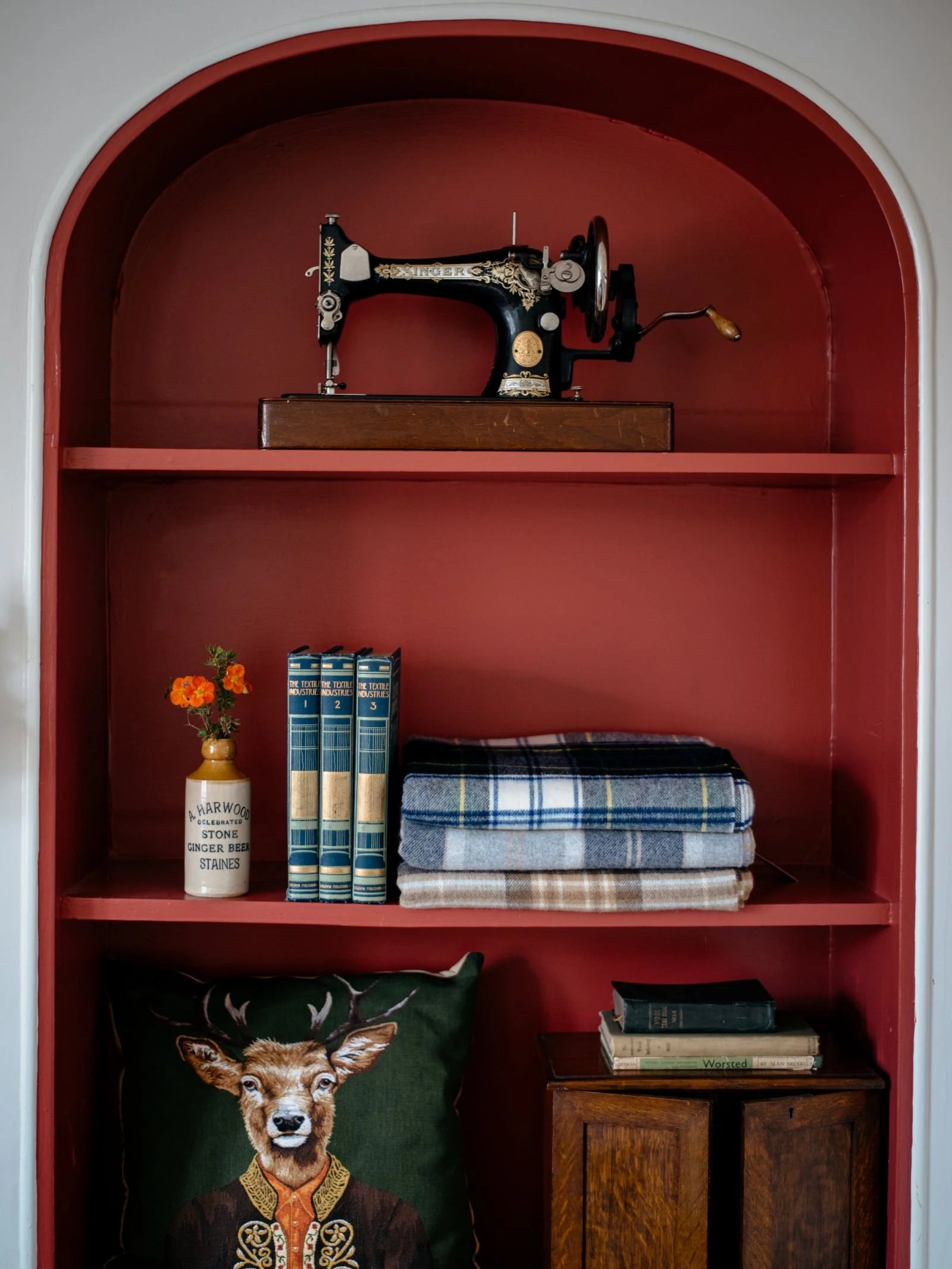A red alcove shelf displays a vintage sewing machine, a vase with orange flowers, books, a Campbell's of Beauly Lambswool Tartan Blanket—including a heritage throw—and a deer-in-clothing pillow. More books and a wooden box sit below.