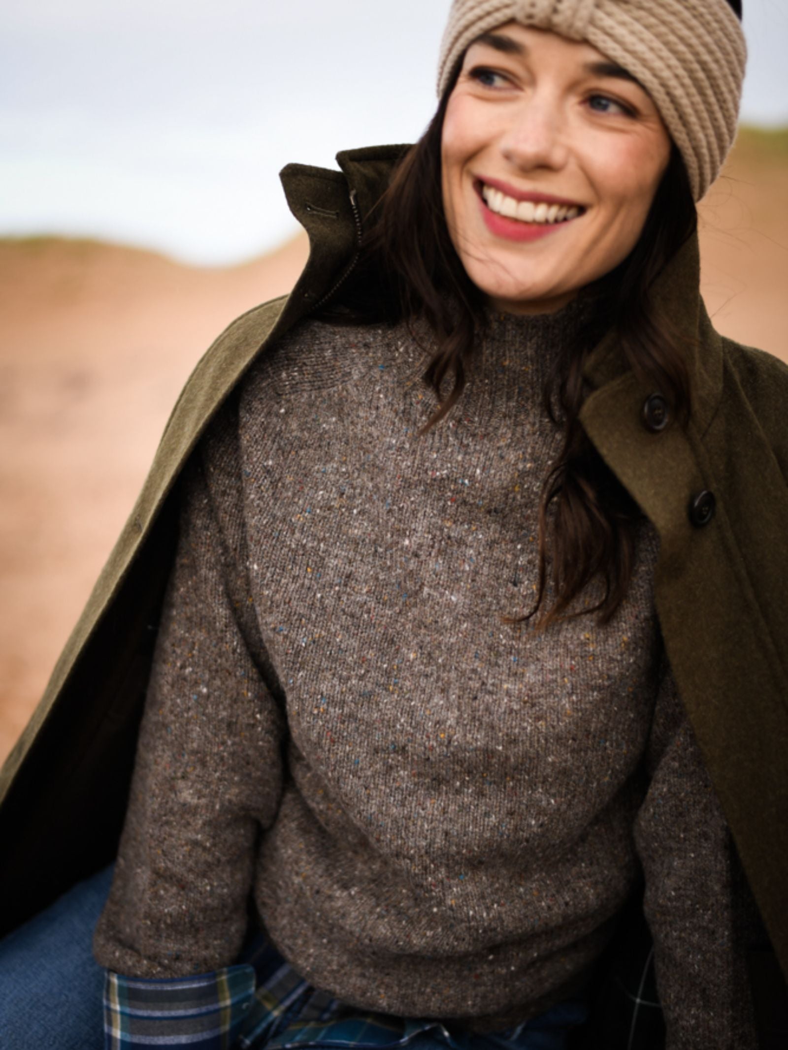 A woman smiles outdoors, wearing a knitted beige headband, a thick brown turtleneck sweater, an olive-green coat draped over her shoulders, and blue jeans. The background is softly blurred.