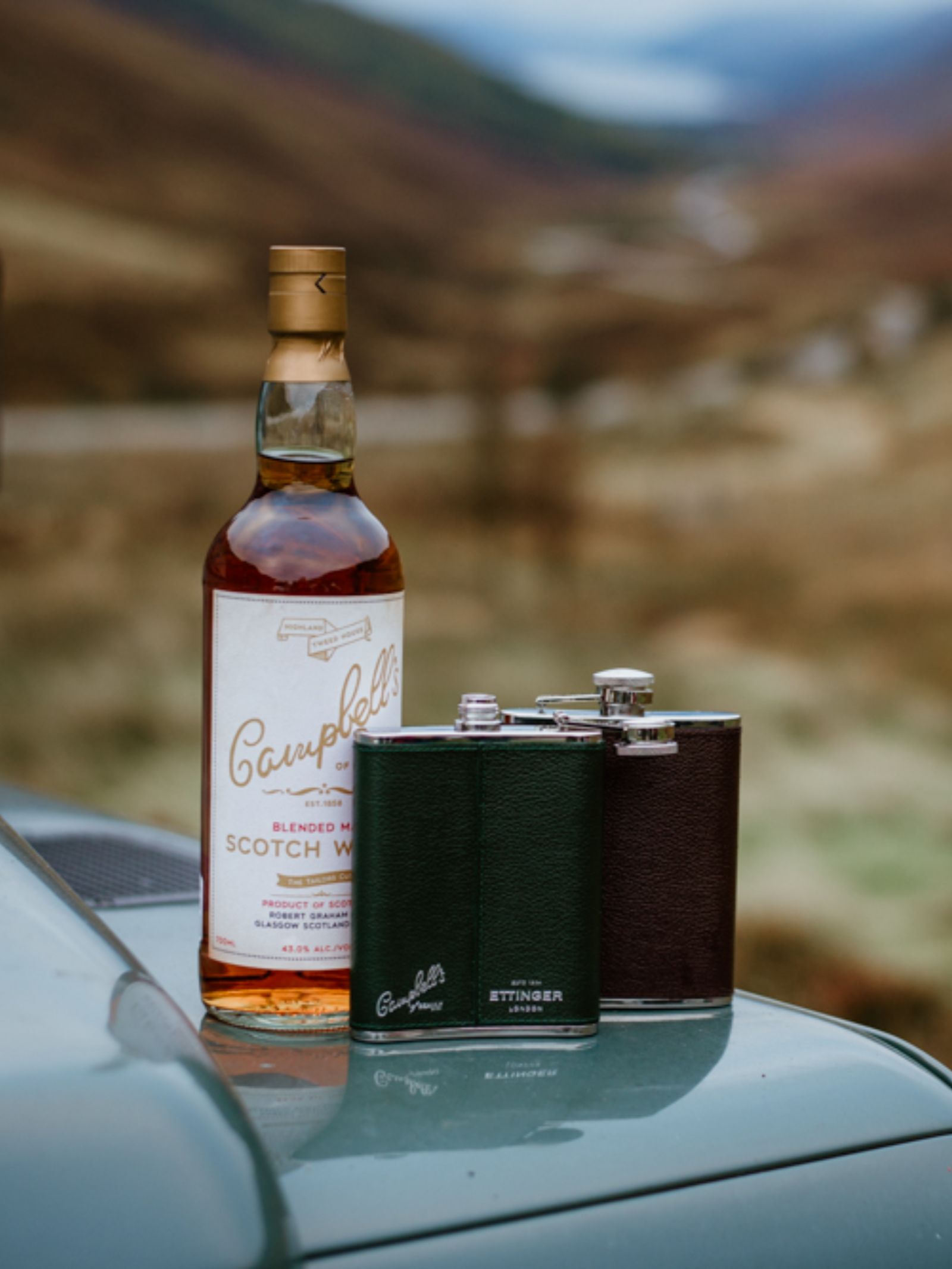 A bottle of Campbell’s Scotch whisky and two 6oz Forest Green Hip Flasks by Campbell's of Beauly x Ettinger rest on a vehicle’s hood, with rolling hills and a winding road blurred in the scenic background.