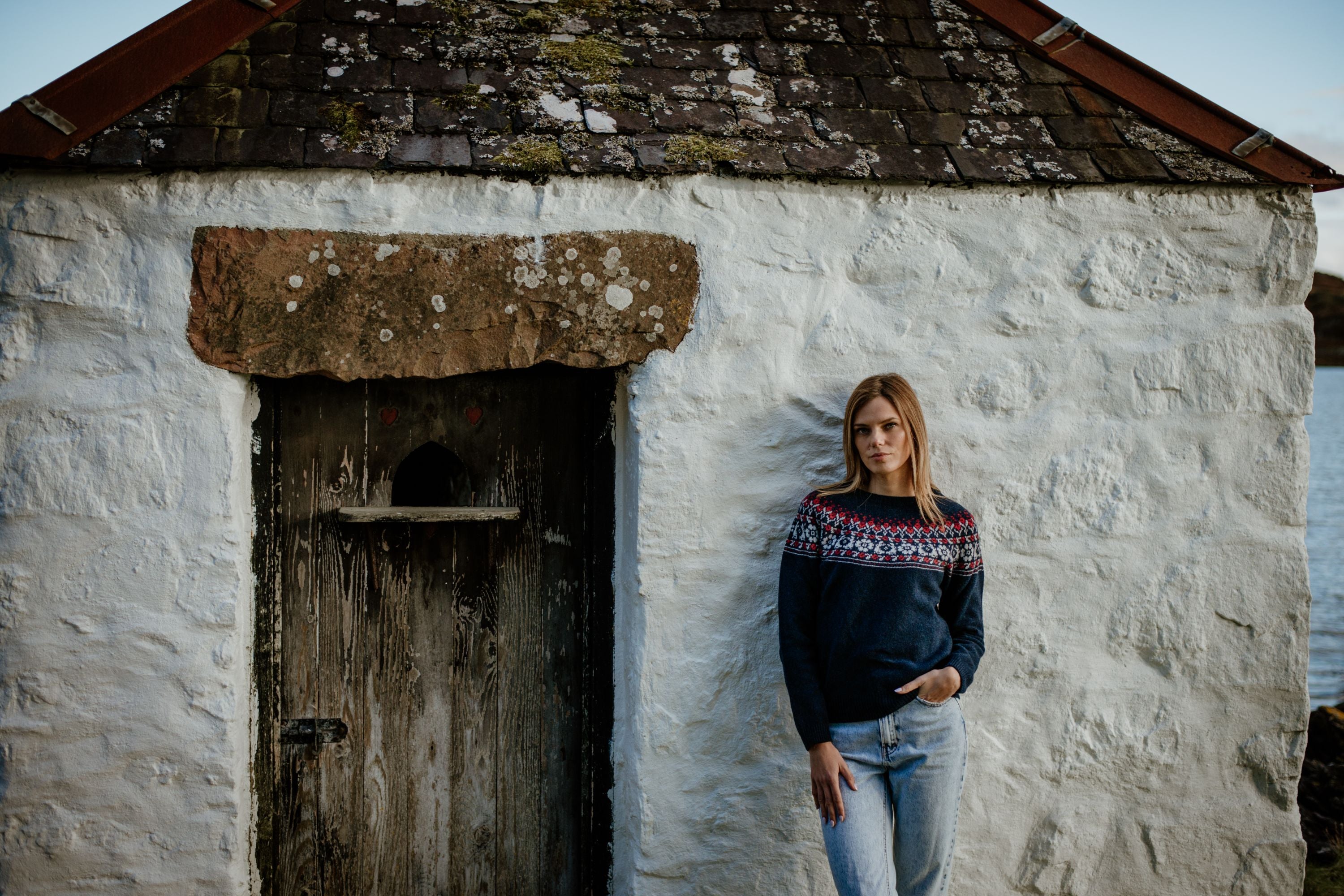 A woman in a blue patterned sweater and jeans stands against the white stone wall of a rustic building with a weathered wooden door and mossy roof, looking at the camera.