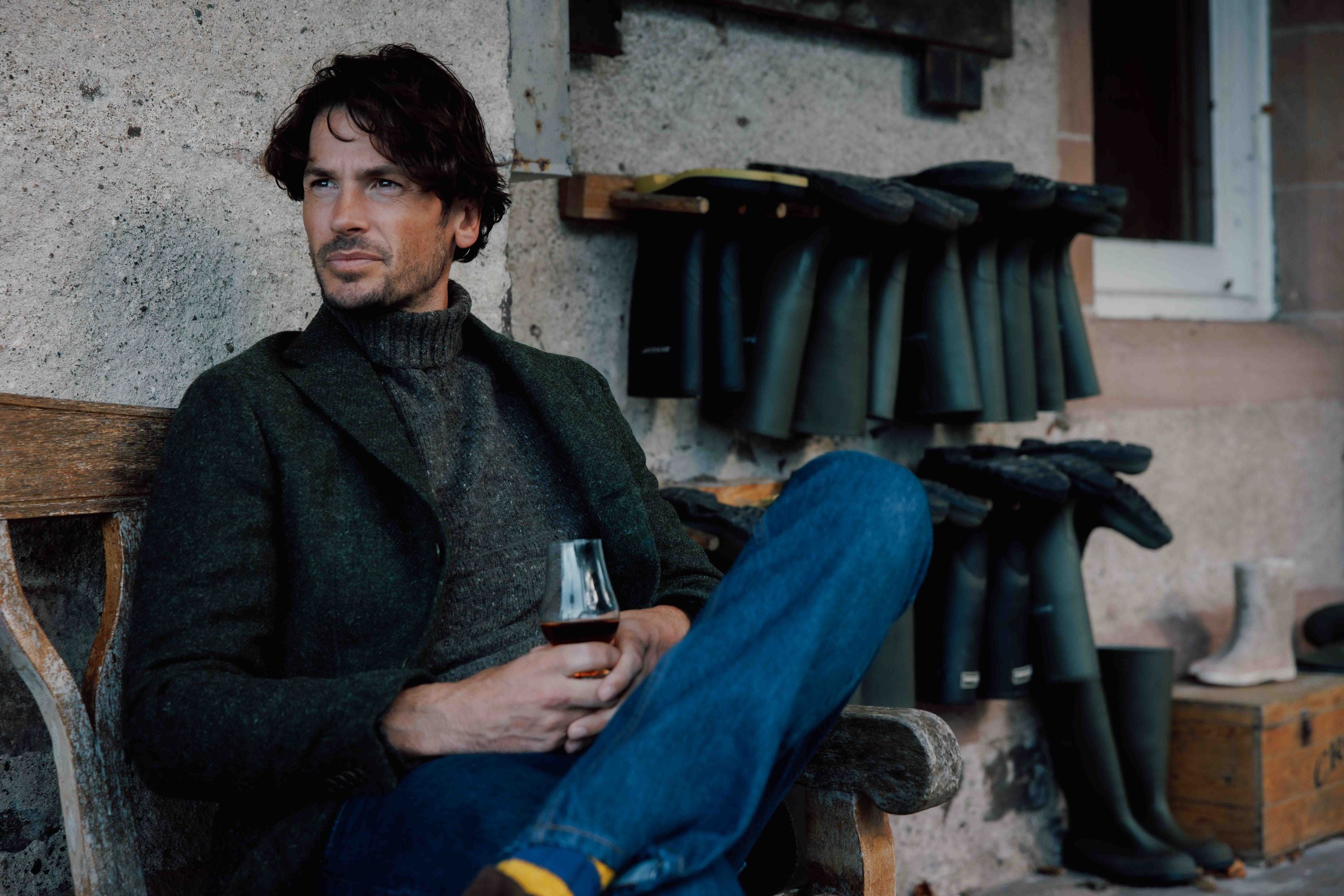 A man in a dark sweater and jacket sits on a wooden bench, holding a glass of wine. He looks to the side thoughtfully. Multiple pairs of rubber boots are lined up on a rack behind him against a stone wall.