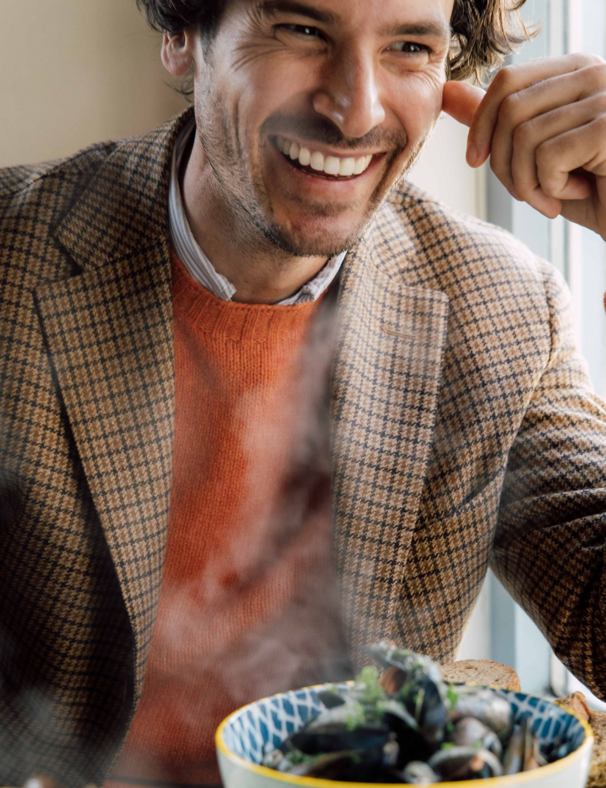 A smiling man wearing a brown checked blazer and orange sweater sits at a table with a steaming bowl of food in front of him.