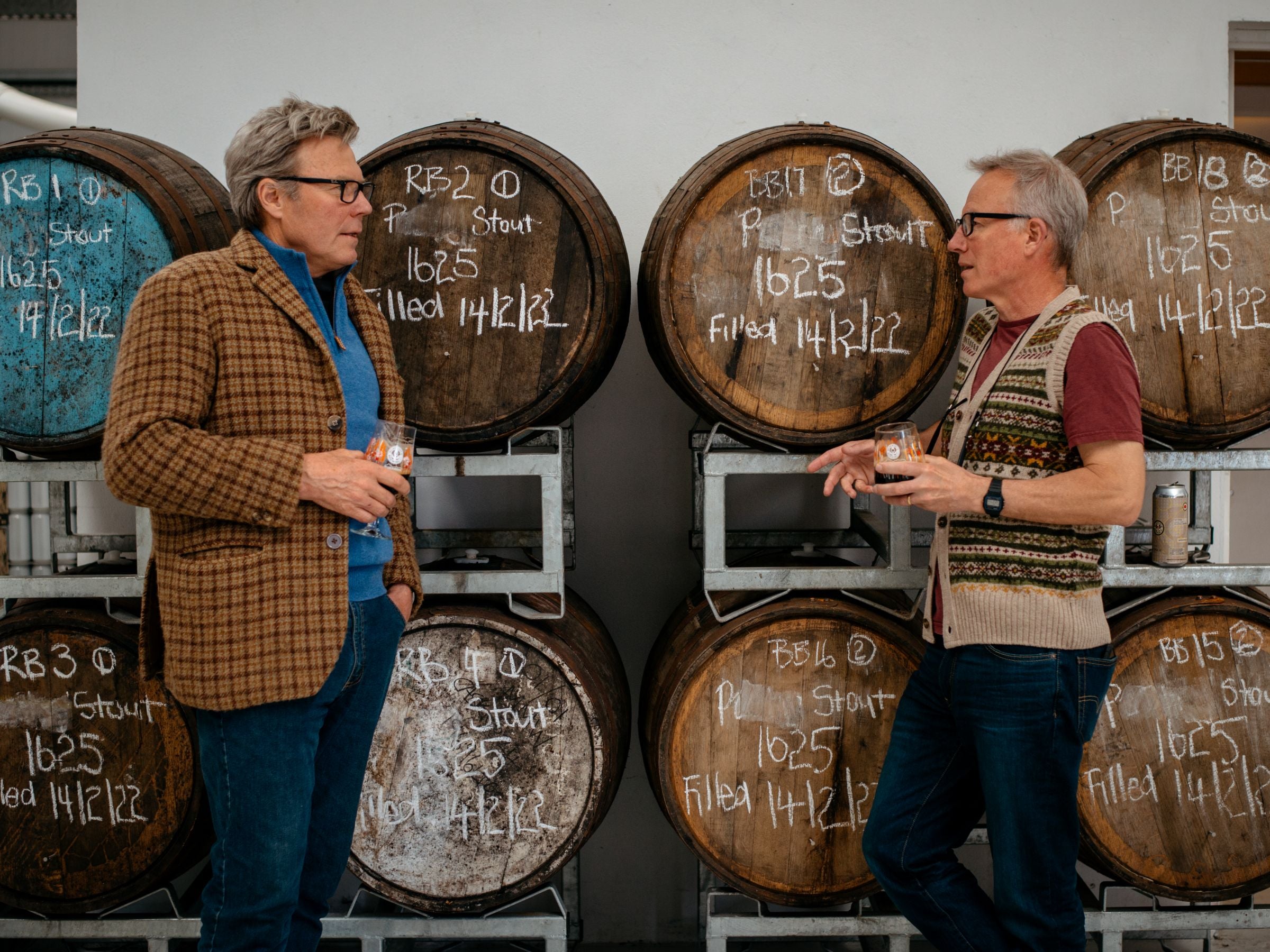 Two men stand and talk while holding drinks in front of large wooden barrels labeled with chalk writing. The barrels are stacked on metal racks in a brewery or distillery setting.