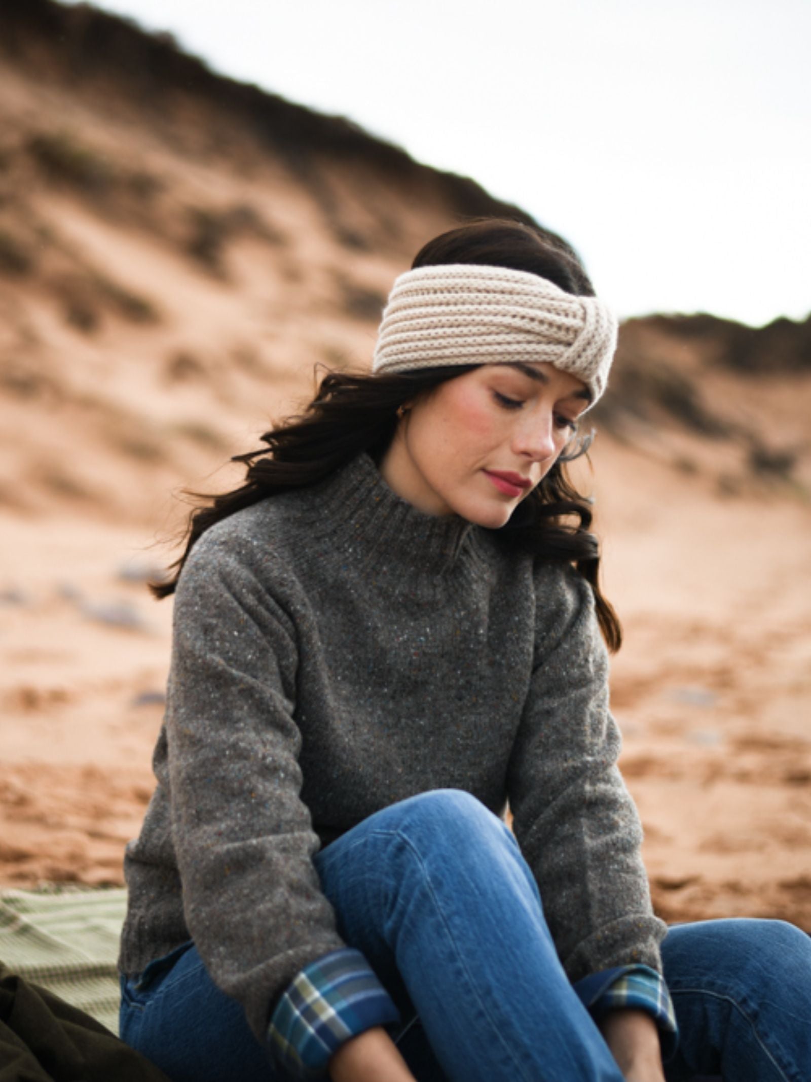 A woman with long dark hair, in a gray sweater and blue jeans, wears the Campbell's of Beauly Cashmere Ear Warmer—a cozy winter accessory—while sitting calmly on a sandy beach with dunes and a cloudy sky in the background.