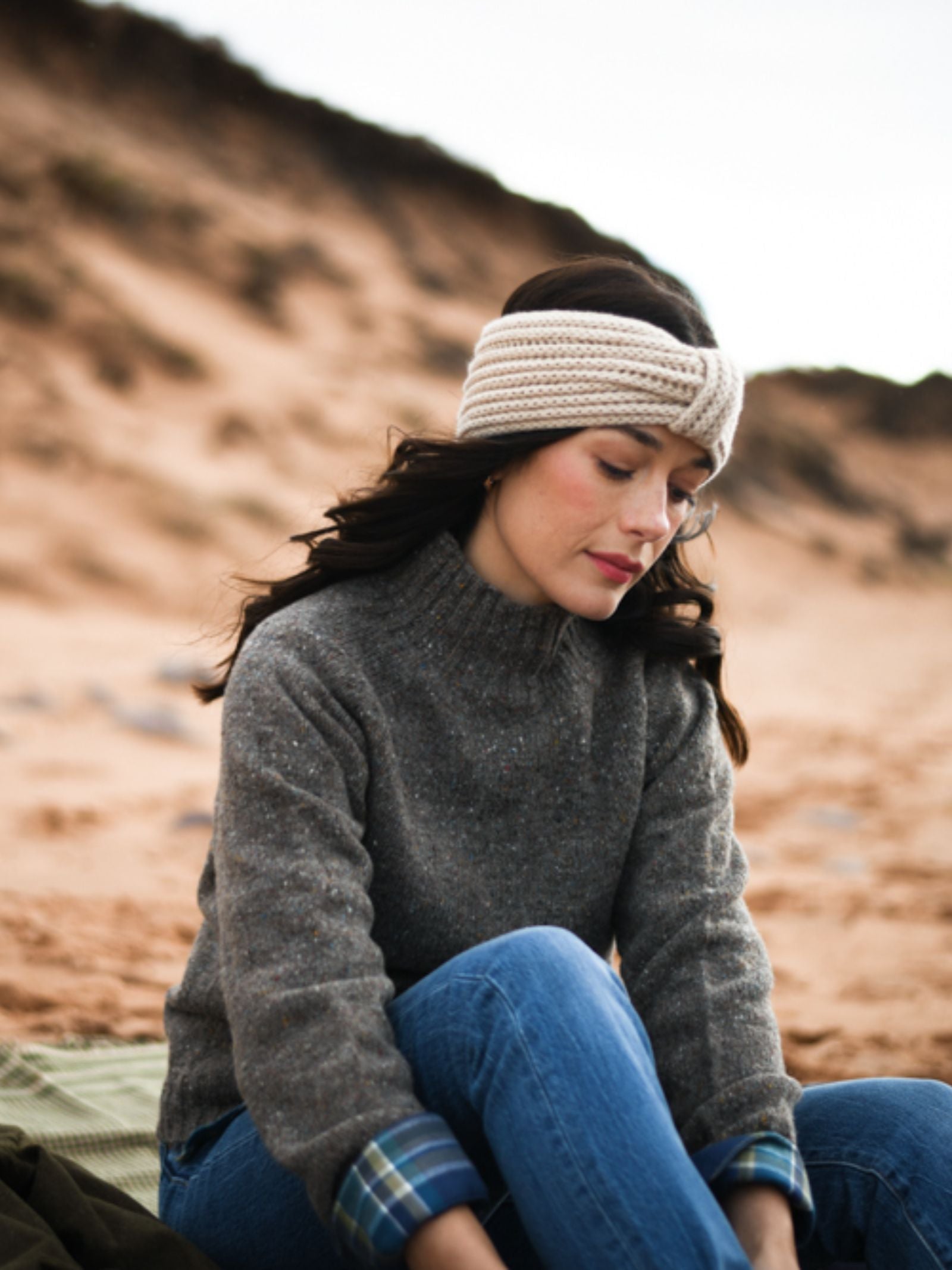 A woman in a gray Campbell's of Beauly Donegal Lambswool Funnel Neck Jumper, blue jeans, and a knitted headband sits on a sandy beach with dunes behind her, gazing down with a calm expression.