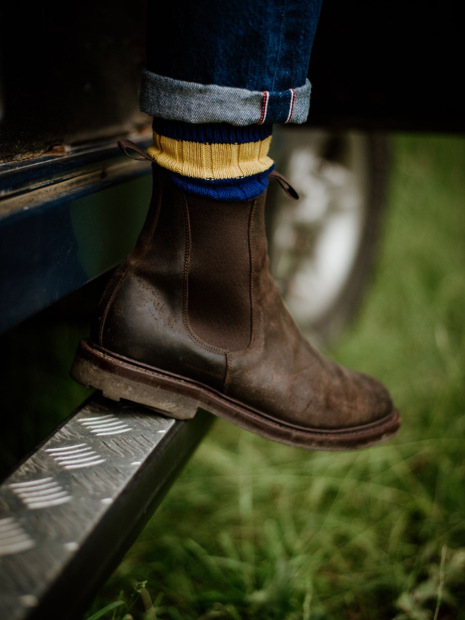 A close-up of a person’s foot in Campbell's of Beauly Lowlander Boots and yellow socks with blue stripes, standing on a metal step near grass, with jeans rolled up at the ankle.