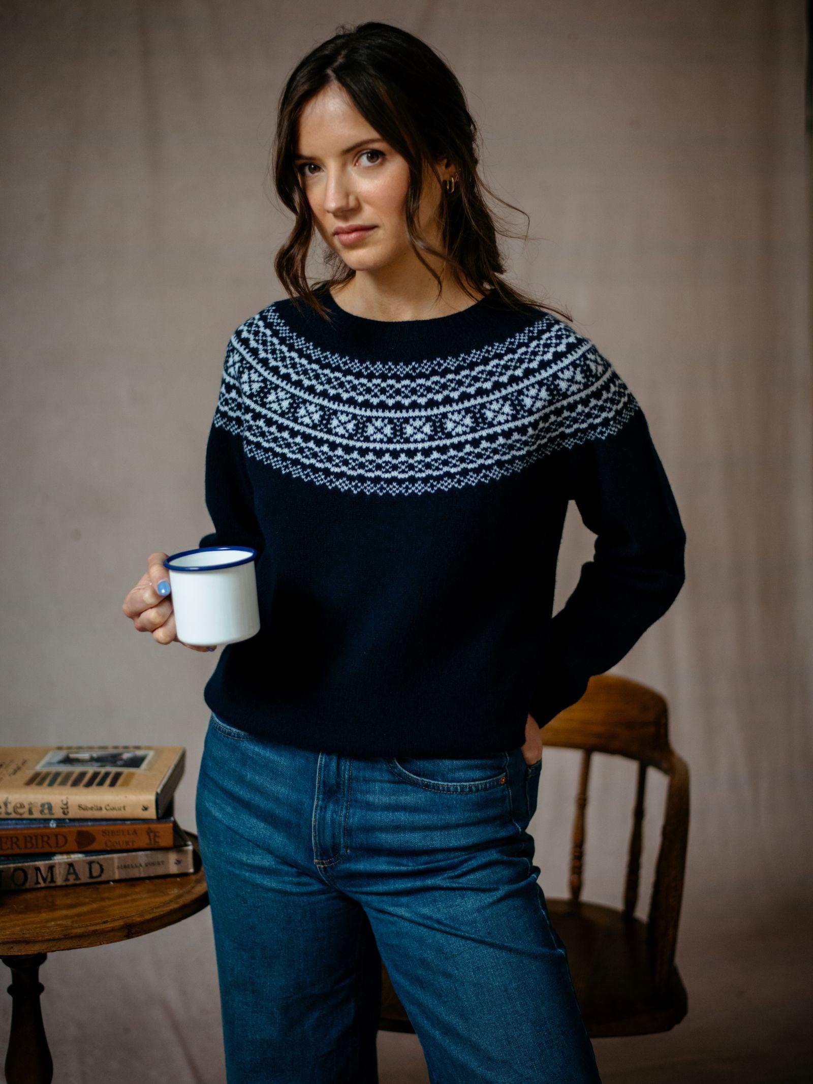A woman with long brown hair wears the Campbell's of Beauly Two-Colour Fairisle Yoke Jumper in navy Geelong lambswool, holding a white mug by a table with stacked books and a wooden chair behind her.