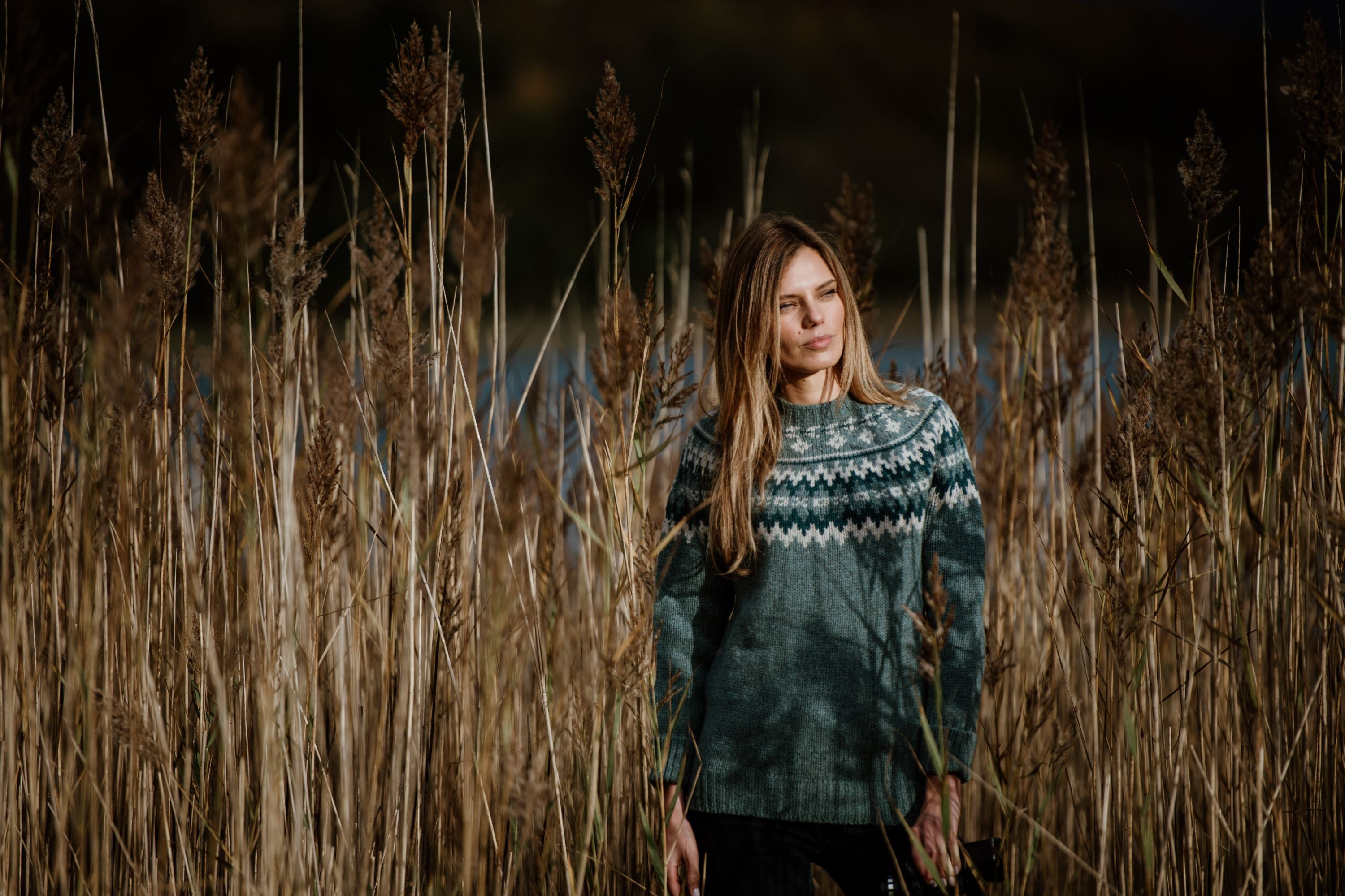 A woman with long hair stands in tall, dry grass, wearing a green patterned sweater. The background is blurred, highlighting the natural outdoor setting.