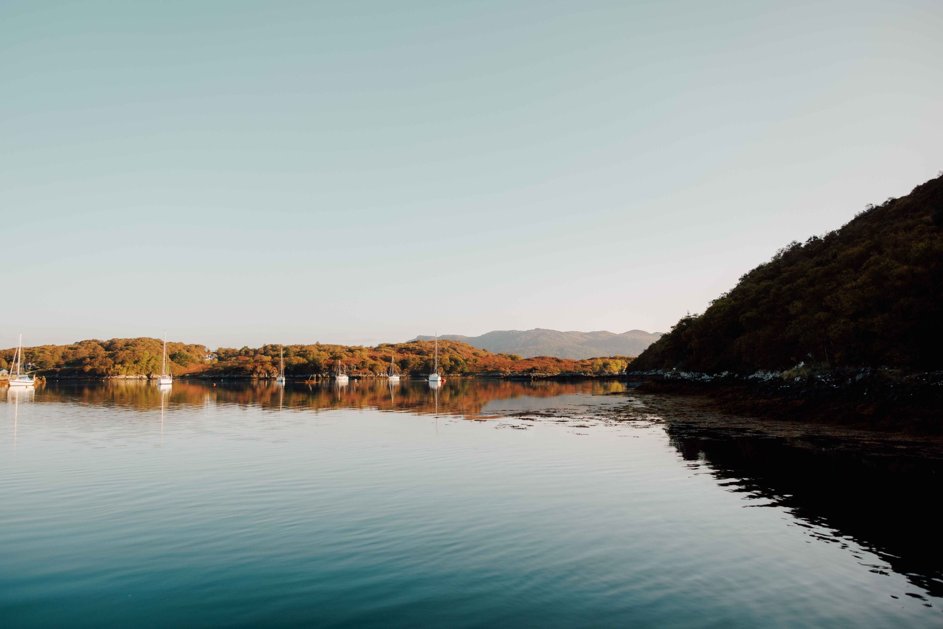 A calm lake reflects the surrounding hills and sky, with a few sailboats anchored near the shore. The landscape features autumn-colored trees beneath a clear, pale sky.