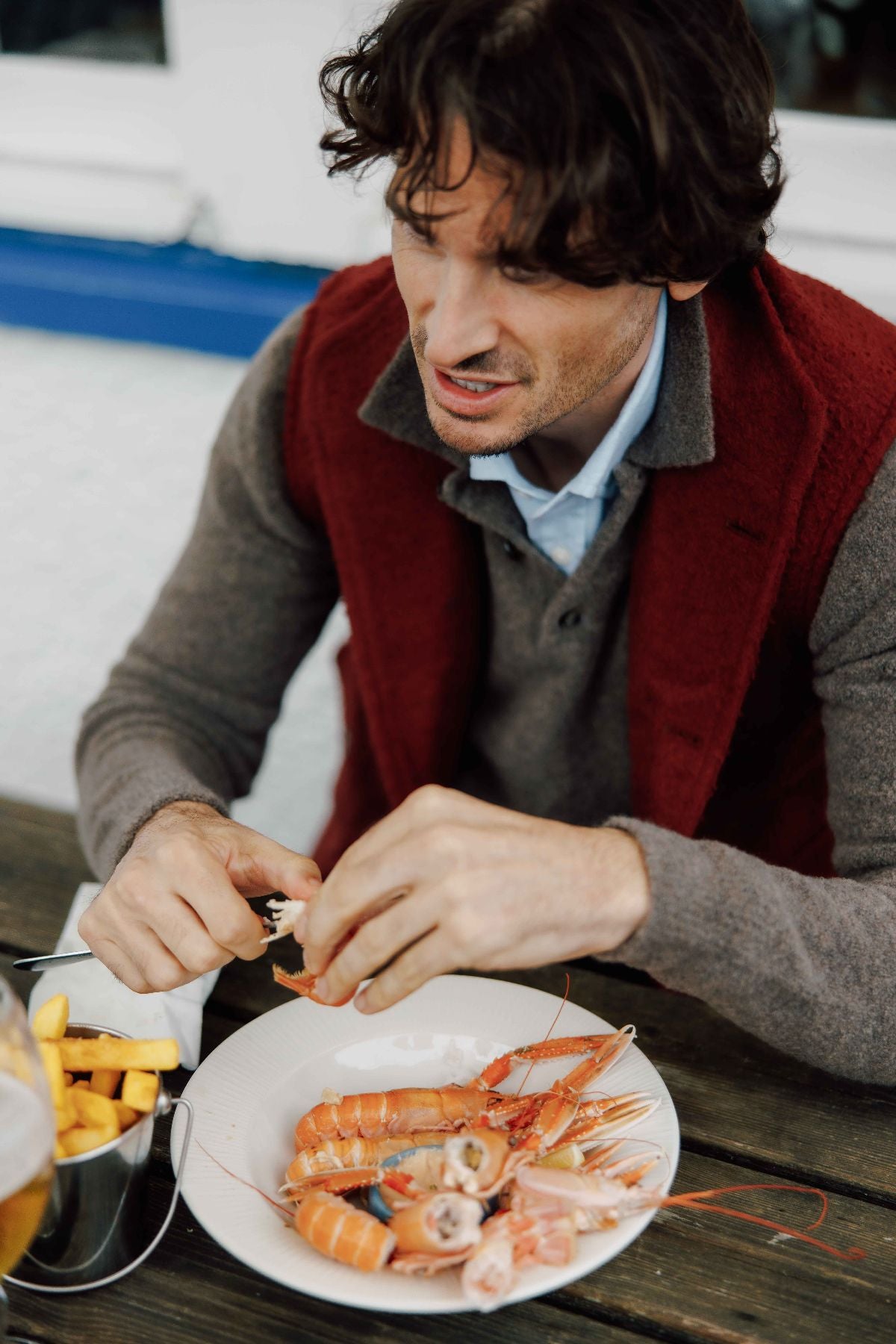 A man with wavy brown hair sits at a table peeling shellfish, with a plate of prawns and a bowl of French fries in front of him. He is wearing a gray sweater and a red vest.