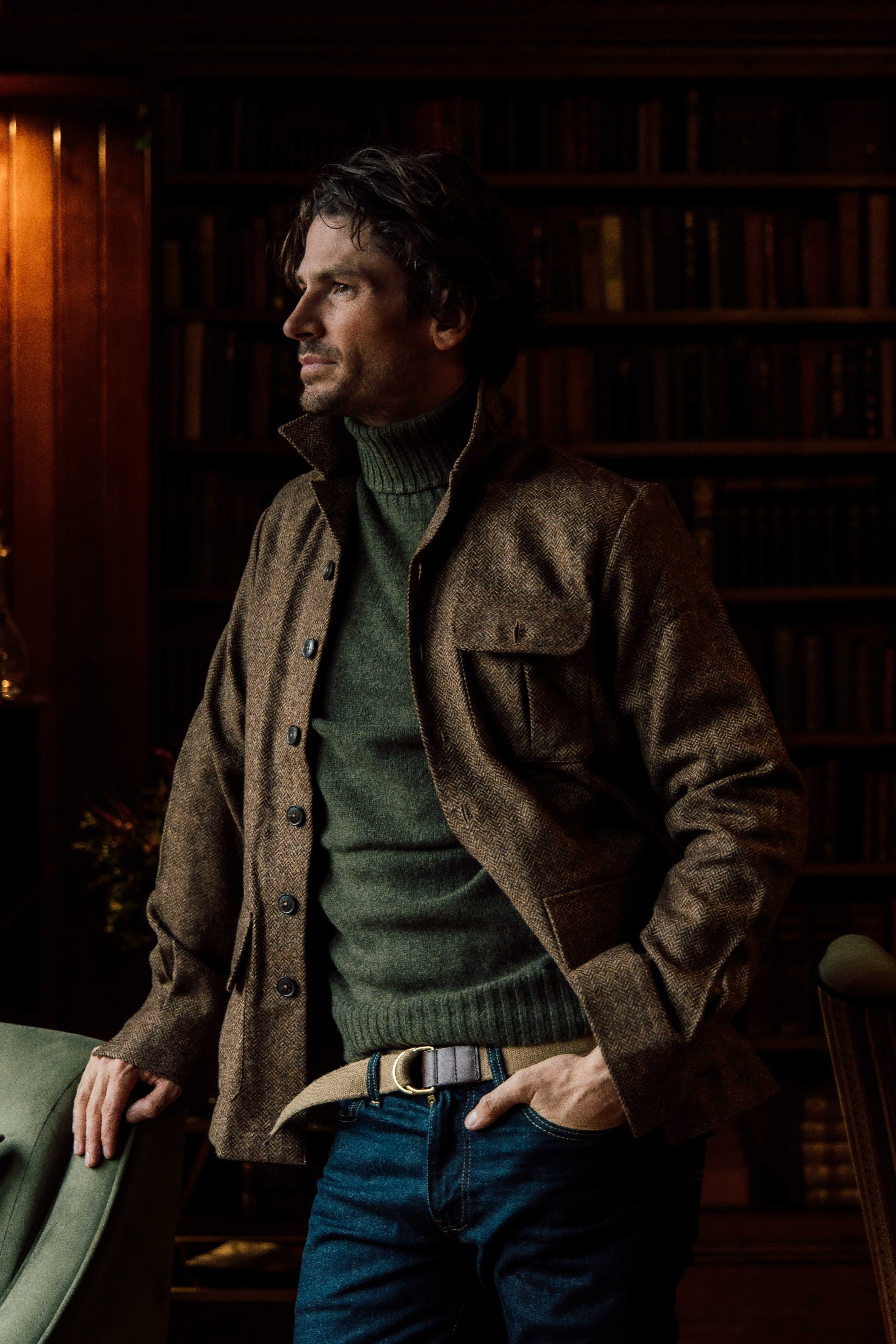 A man with wavy hair stands indoors, wearing a brown jacket over a green turtleneck sweater and jeans. He is leaning on a green chair, with a bookshelf filled with books in the background.