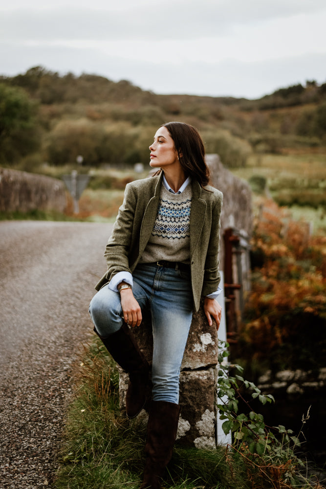 A woman in a green blazer, patterned sweater, jeans, and knee-high boots sits on a stone post beside a rural road, looking to the side. Behind her are green fields and trees under a cloudy sky.