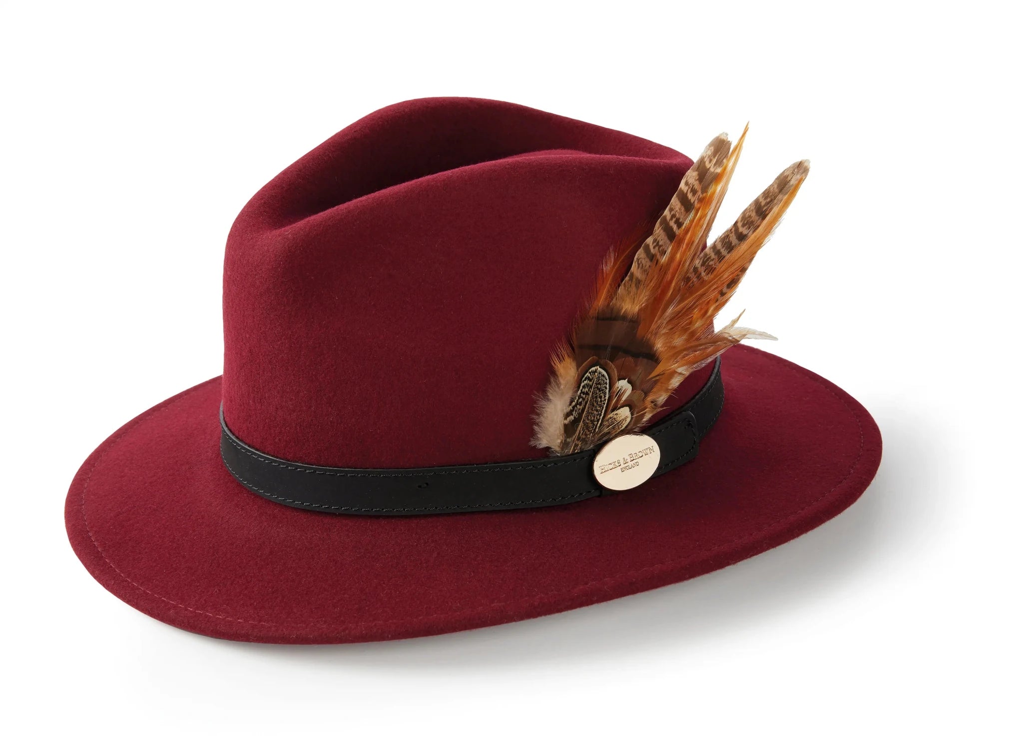 The Hicks & Brown Auld Stock Gamebird Fedora is a burgundy wool felt hat with a black band, feather wrap in brown and beige, and a small round gold pin on the side, shown against a white background.