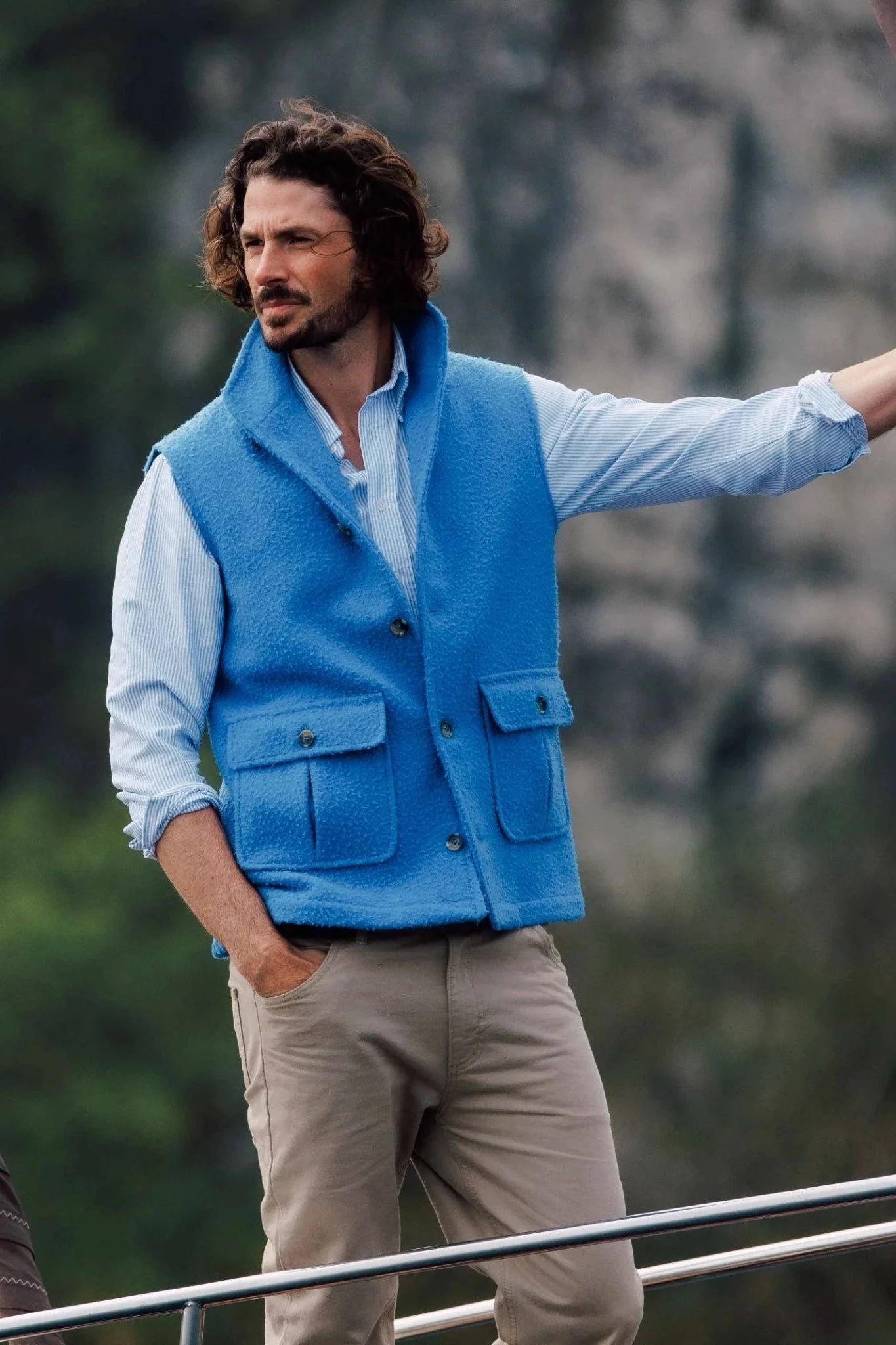 A man with curly hair in a light blue shirt, beige pants, and a bright blue Auld Stock Casentino Fleece Gilet by Campbell's of Beauly stands outdoors beside a railing, with blurred greenery and rocks in the background.