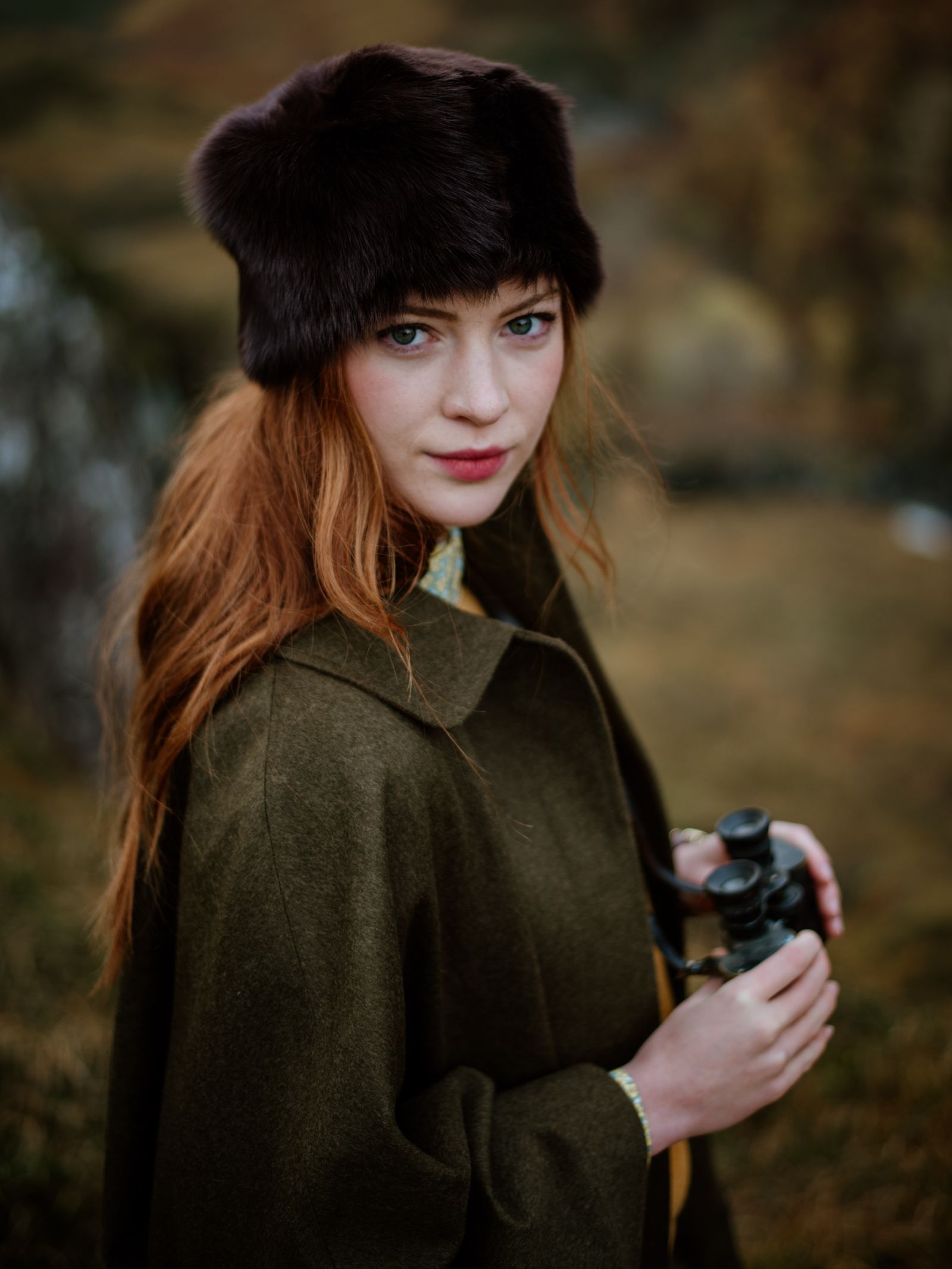 A woman with long red hair wears a dark fur hat and the Loden Green Cape by Campbell's of Beauly while standing outdoors with binoculars, set against a blurred, natural rural landscape.