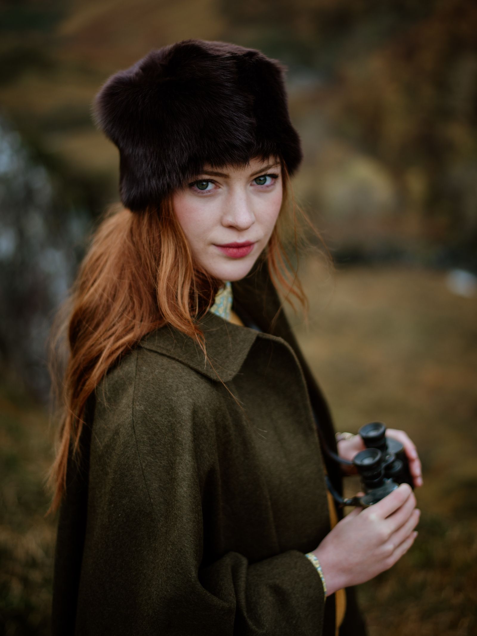 A woman with long red hair wears the Campbell's of Beauly Pillar Box Hat and an olive green coat, holding binoculars outdoors in an autumnal landscape, looking directly at the camera.