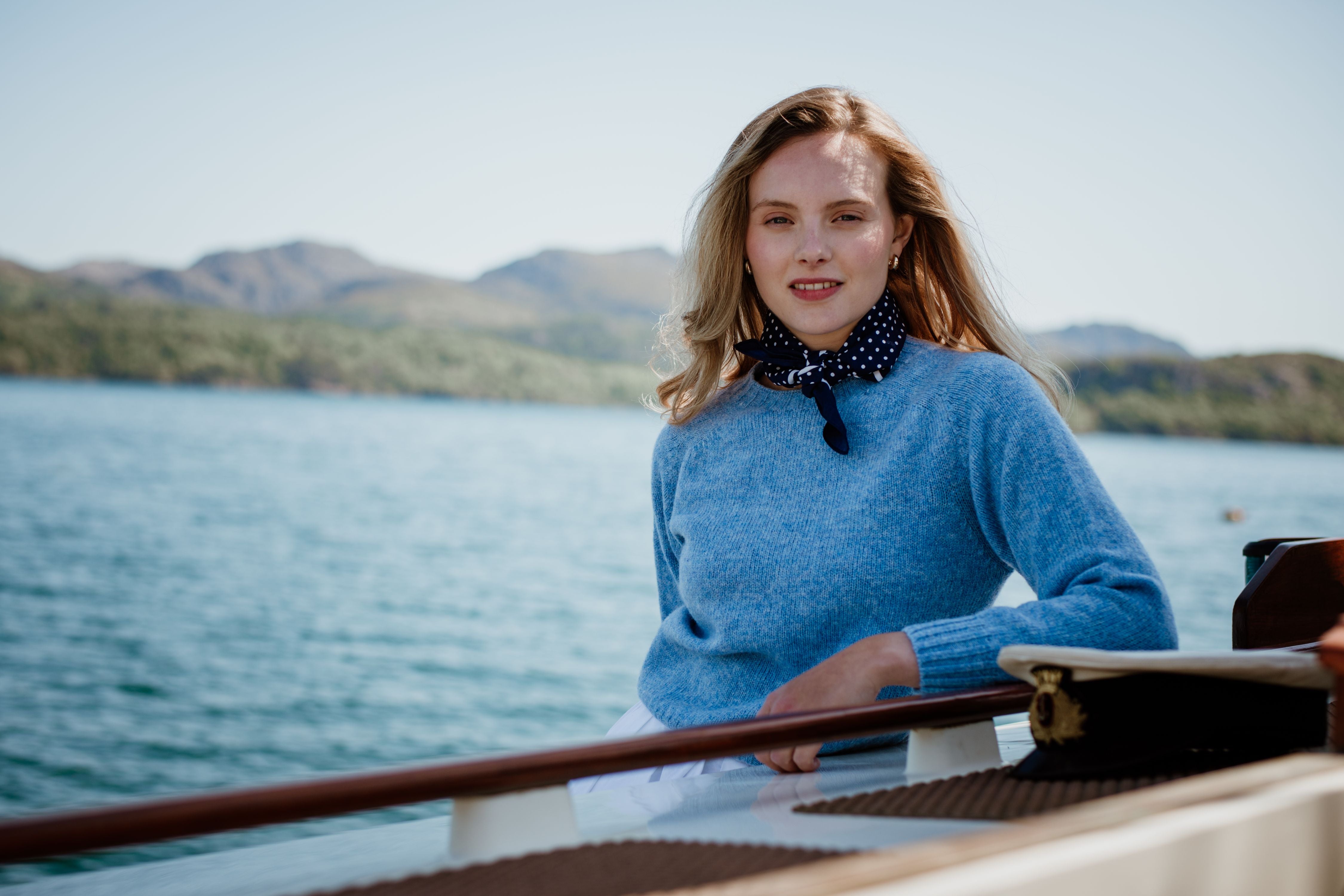 A woman with blonde hair wearing a blue sweater and a navy polka dot scarf poses on a boat, with calm blue water and distant tree-covered hills in the background on a sunny day.