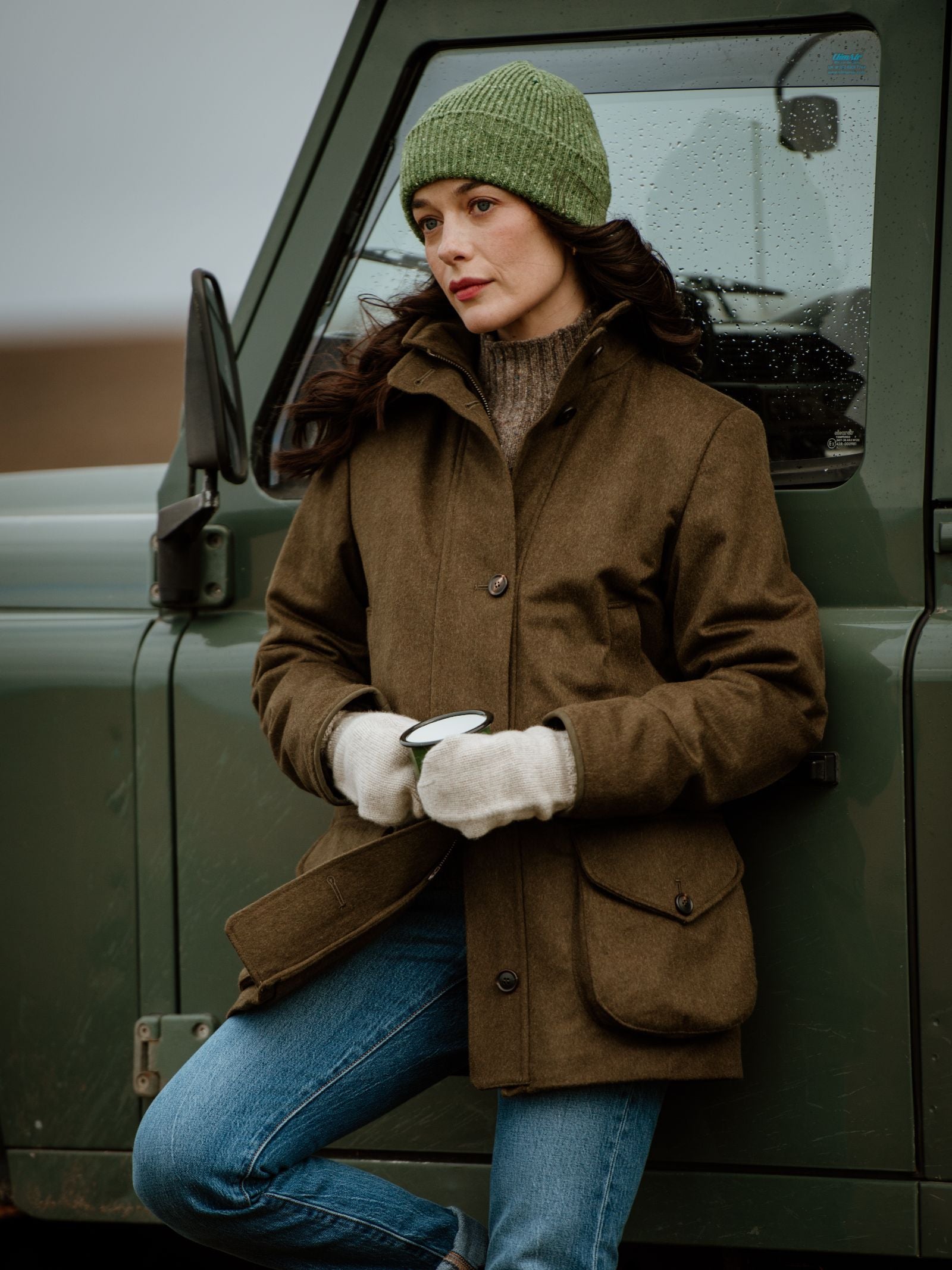 A woman wearing a green Campbell's of Beauly Donegal Beanie, brown jacket, jeans, and white gloves leans against a green vehicle outdoors on a cloudy day, holding a cup with a thoughtful expression.