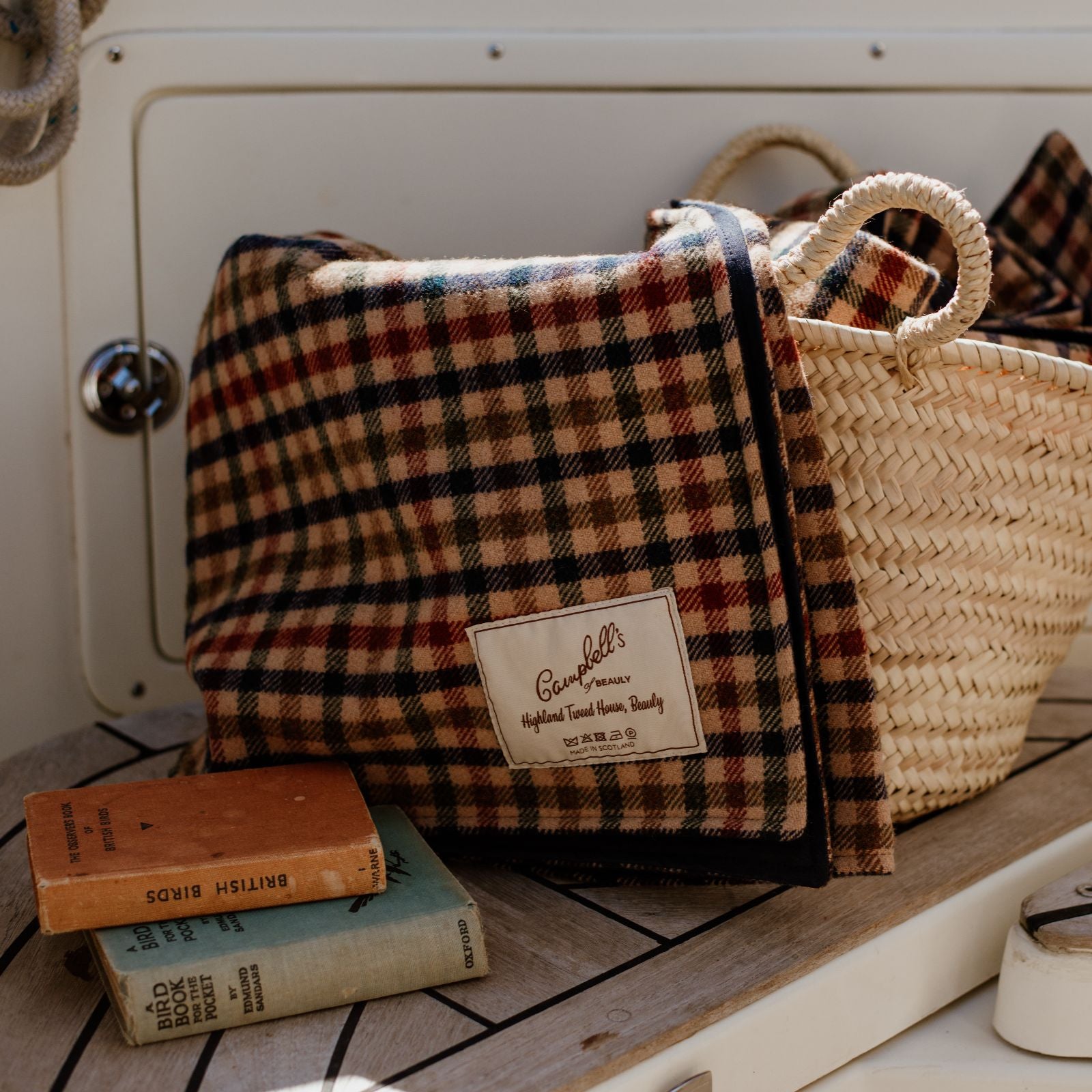 A plaid blanket draped over a wicker basket sits on a wooden surface next to three vintage books. The blanket label reads Campbells Highland Tweed, Luxury Quality.