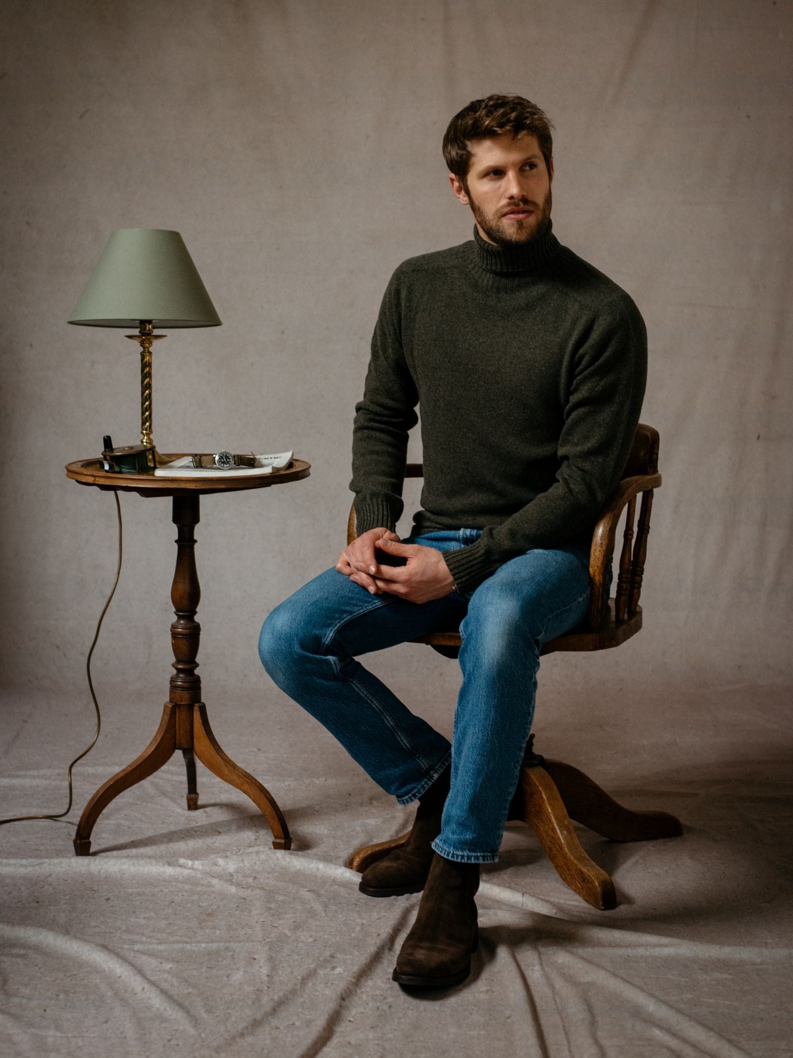 A man with a beard wears the Campbell's of Beauly Lambswool Roll Neck in dark green, blue jeans, and brown boots as he sits on a wooden chair beside a table with a lamp, set against a neutral textured background.